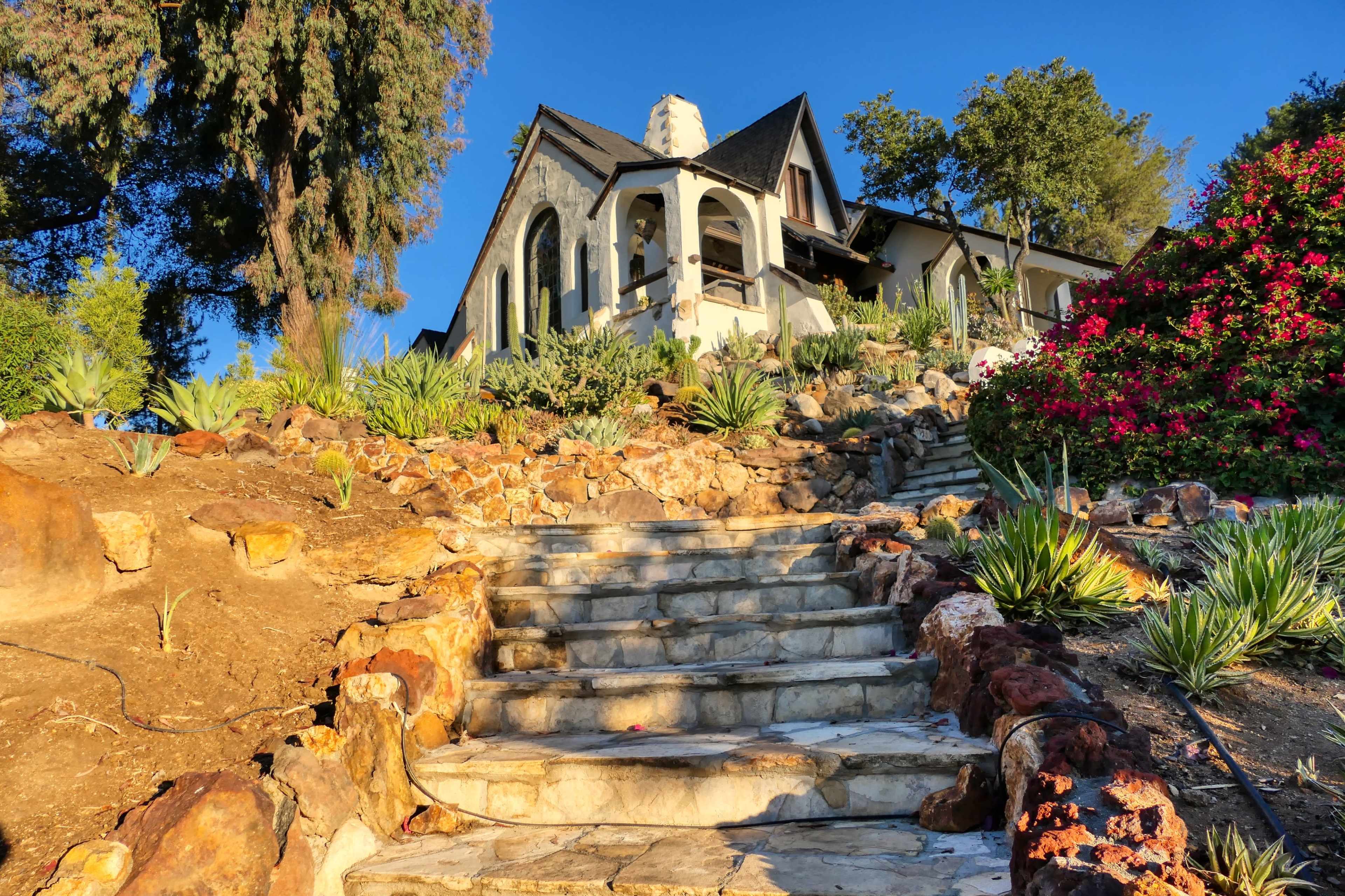 A stone staircase leads up to a large house surrounded by greenery and flowering plants.