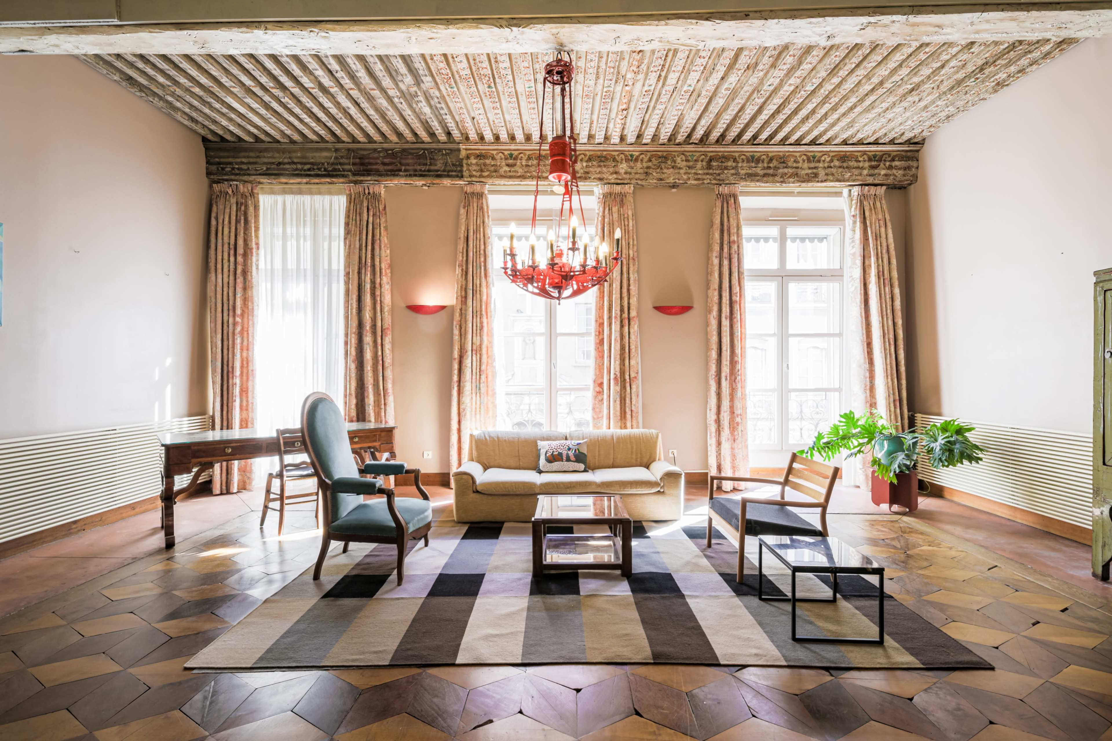 The image shows a spacious living room with a wooden ceiling, large windows, a chandelier, and a mix of wooden and upholstered furniture arranged around a patterned area rug.