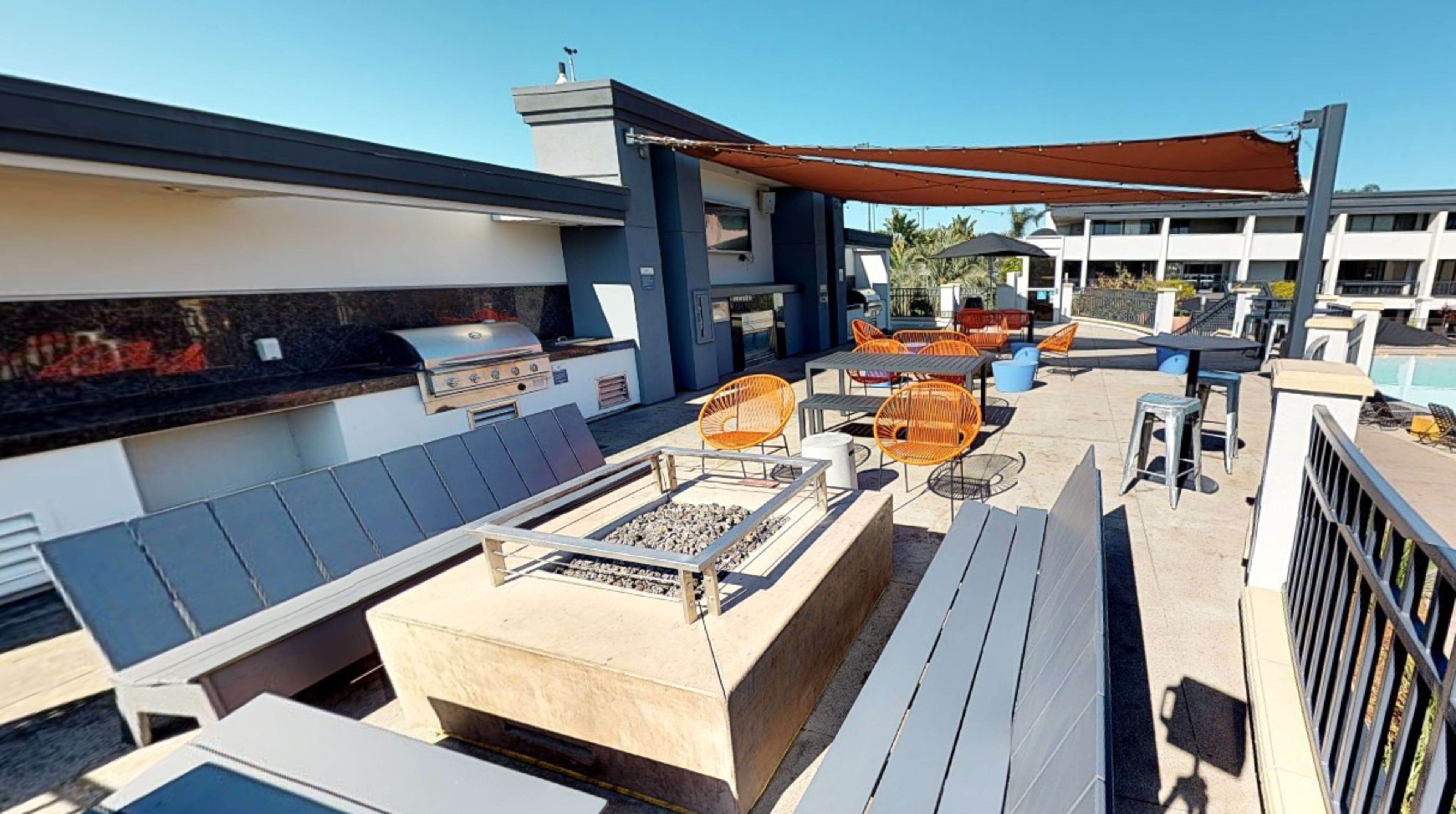 A rooftop terrace with outdoor furniture, a grill station, and a fire pit area, surrounded by a clear blue sky.