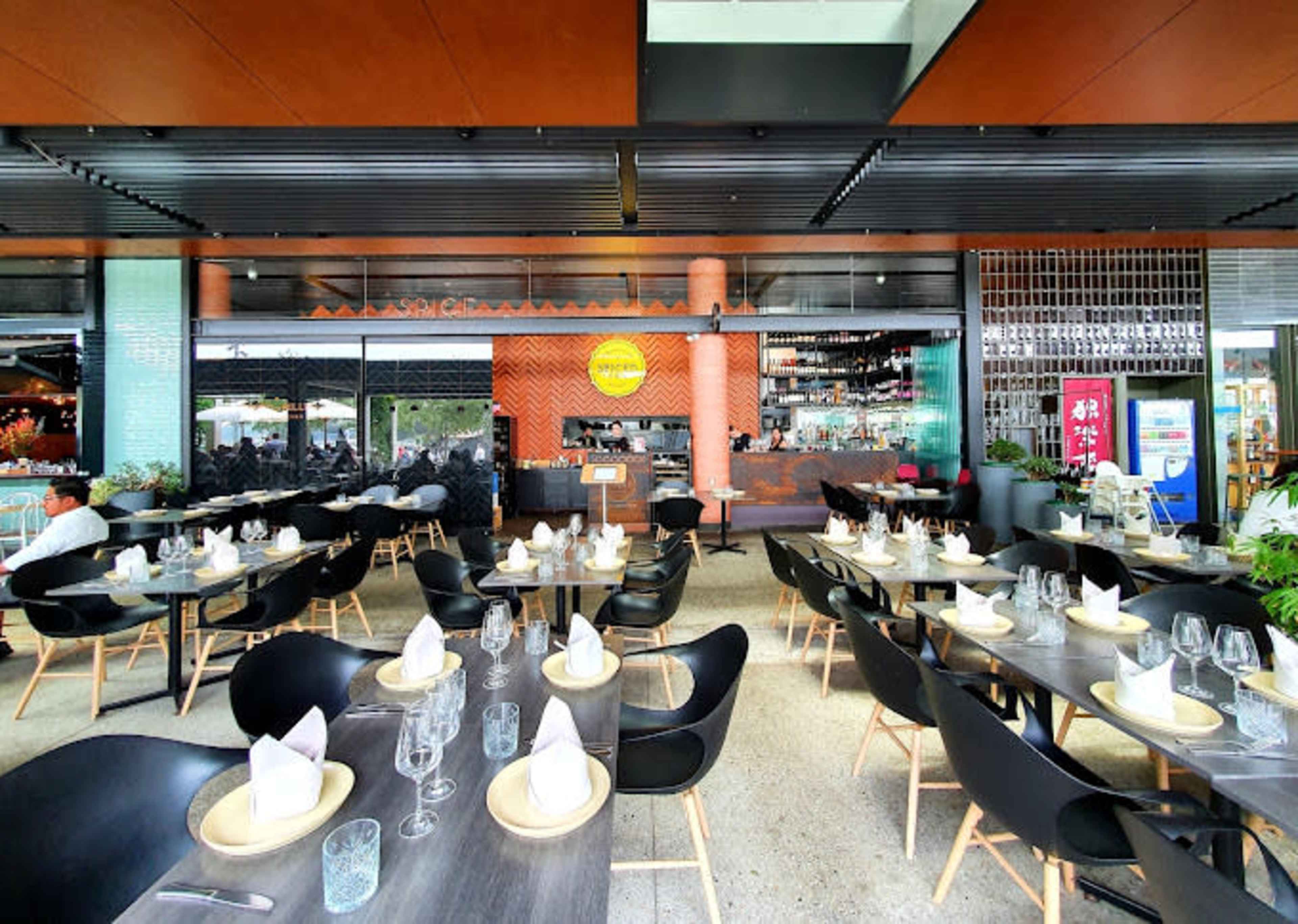 The image shows a modern restaurant exterior with neatly arranged tables and chairs on a patio, and a bar area visible in the background.