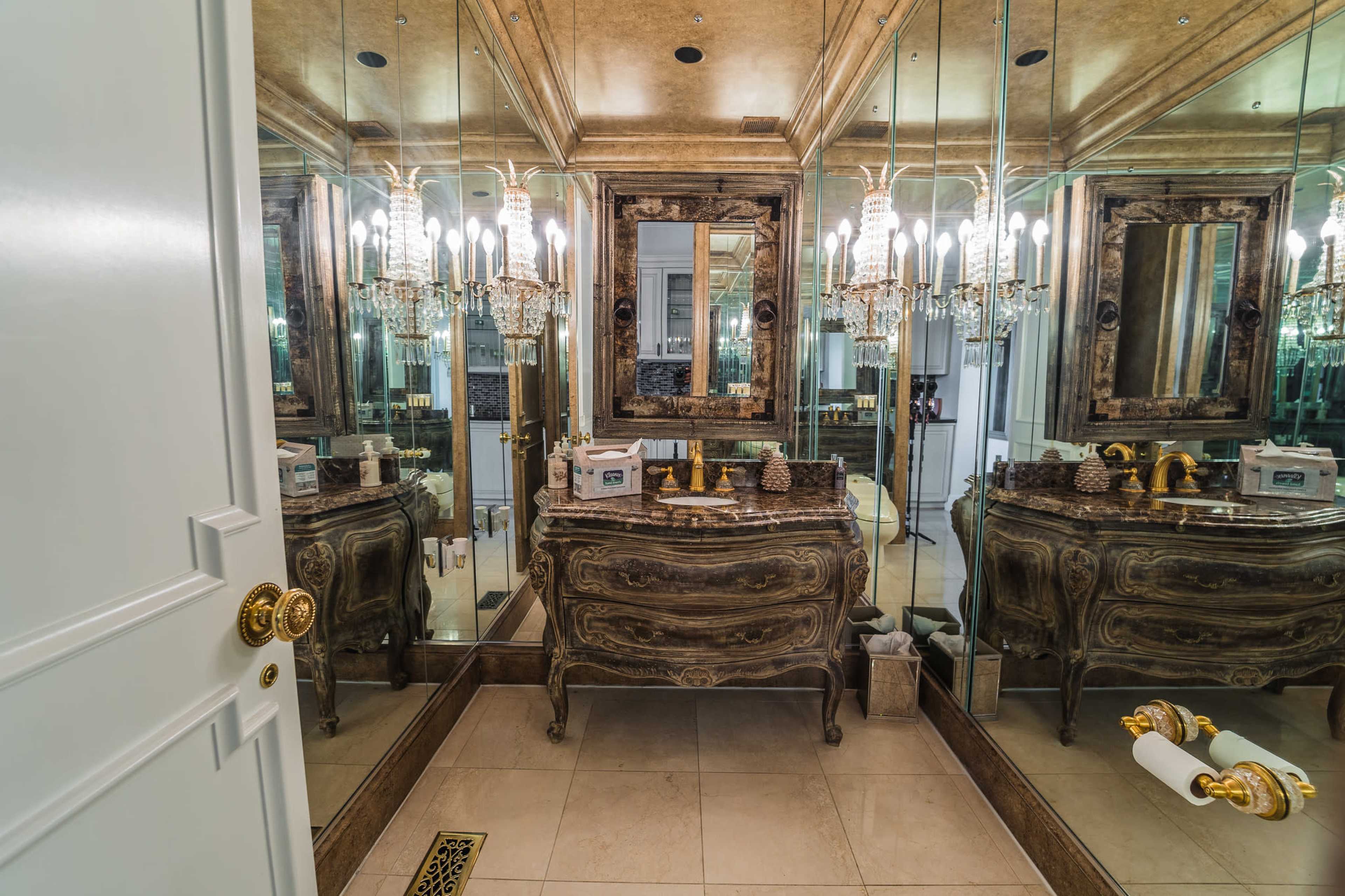A luxurious bathroom features a vintage wooden vanity with ornate detailing, surrounded by mirrored walls and illuminated by multiple chandeliers.