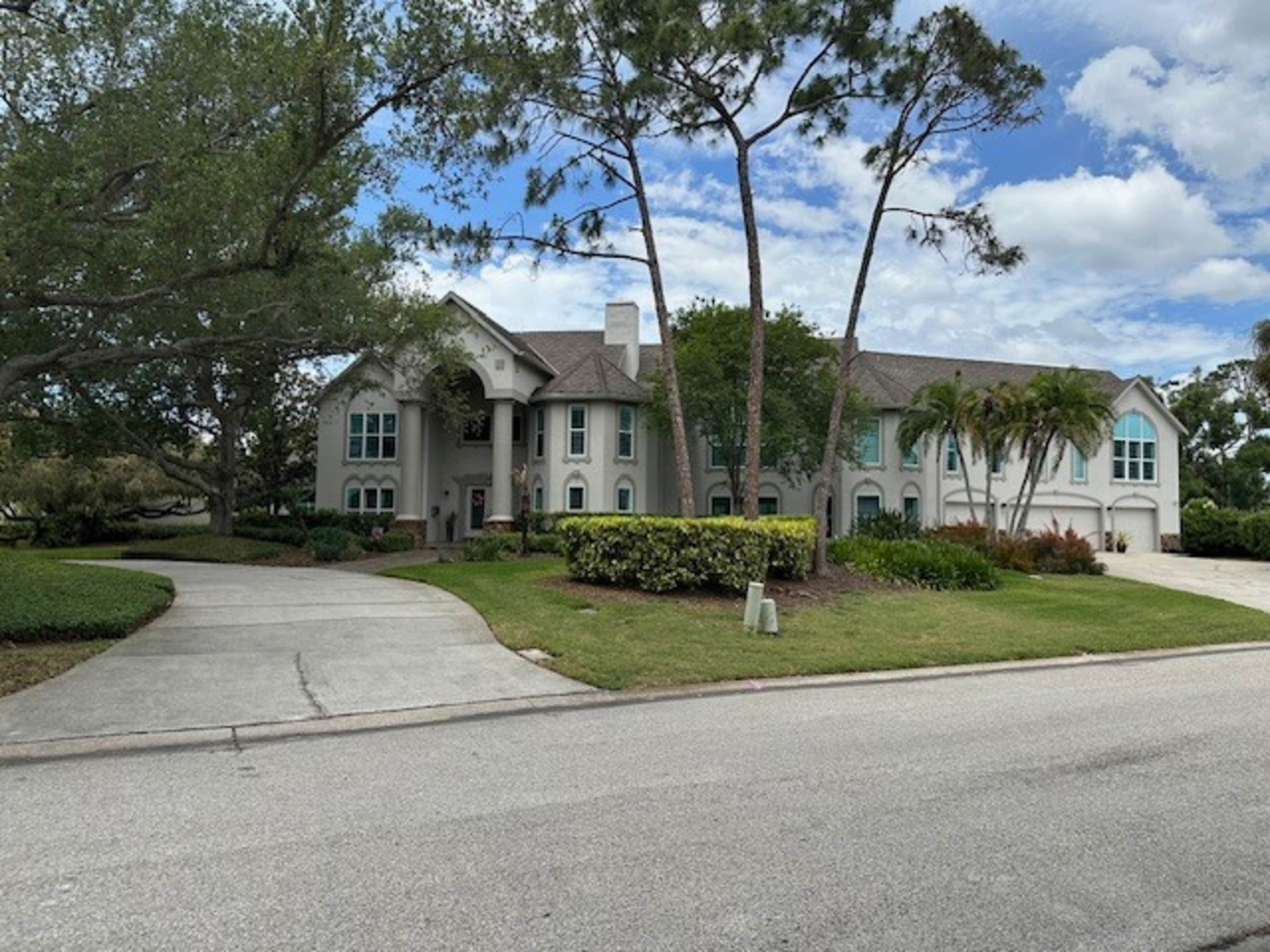 A large, two-story house with a broad driveway is surrounded by trees and landscaping.