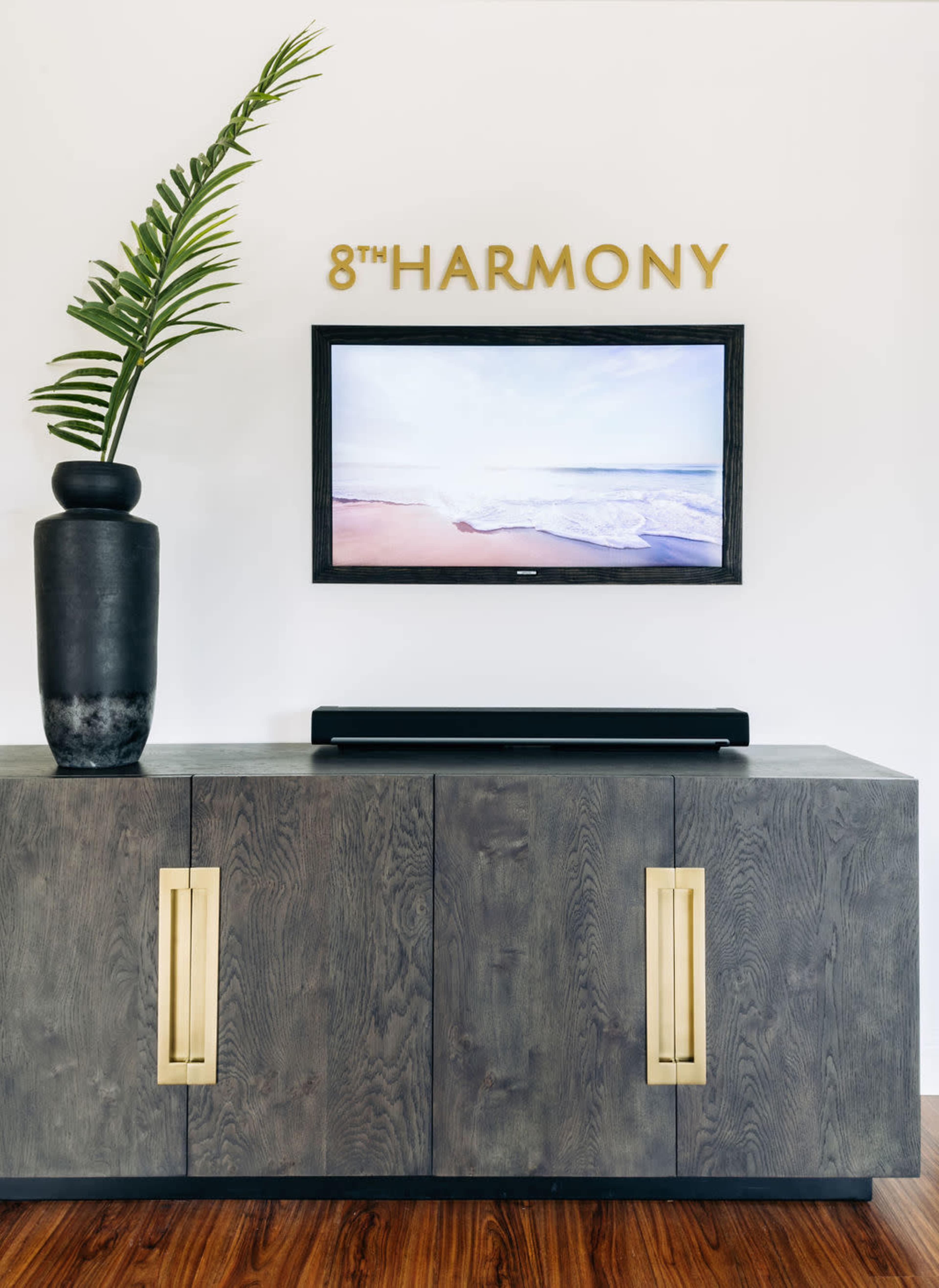 A modern living space features a dark wooden cabinet with gold handles, a tall black vase with a green plant, and a wall-mounted television displaying a beach scene above the cabinet.