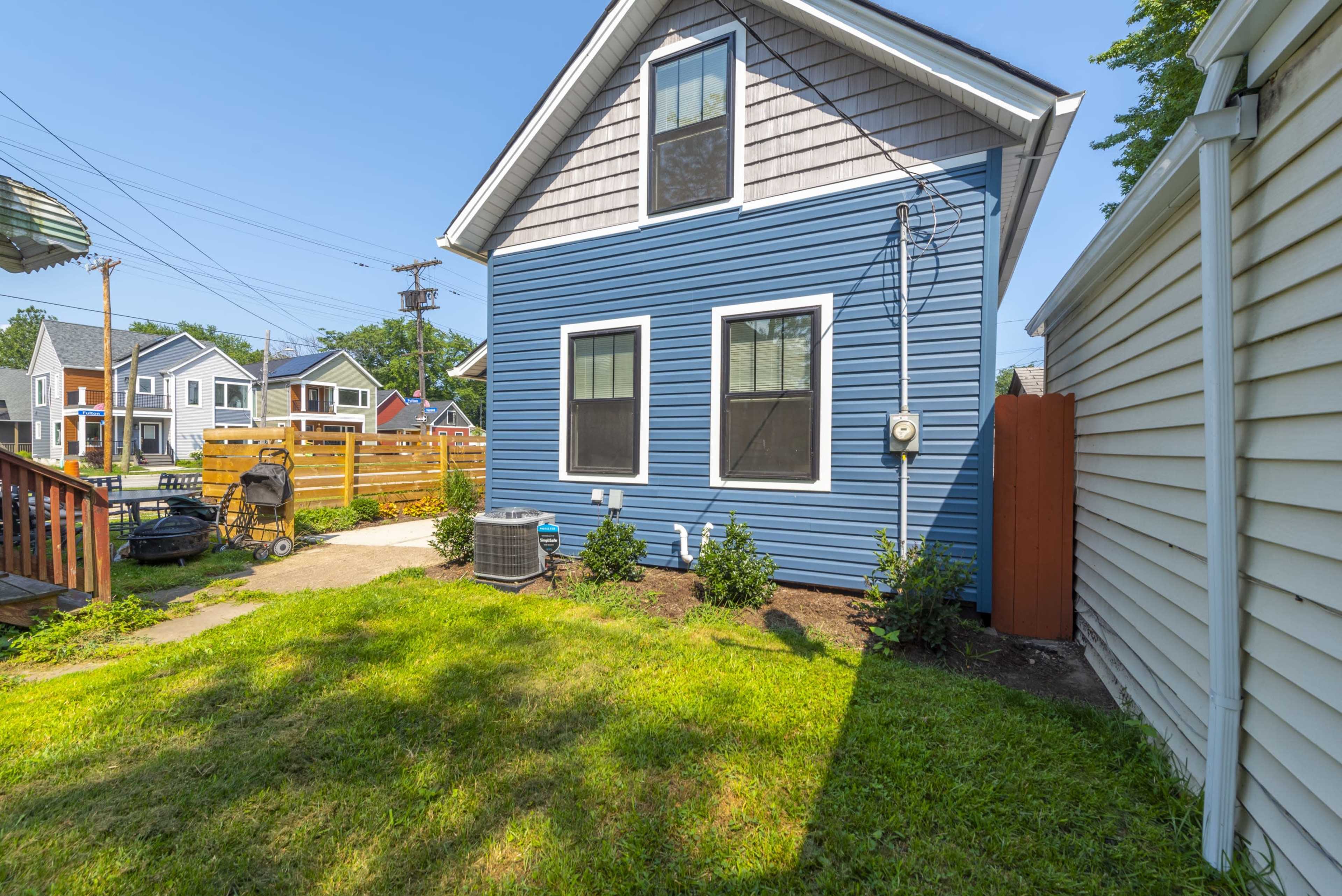 The side yard of a blue house with a garden path, an air conditioning unit, and green grass, bordered by a fence and a neighboring white structure.