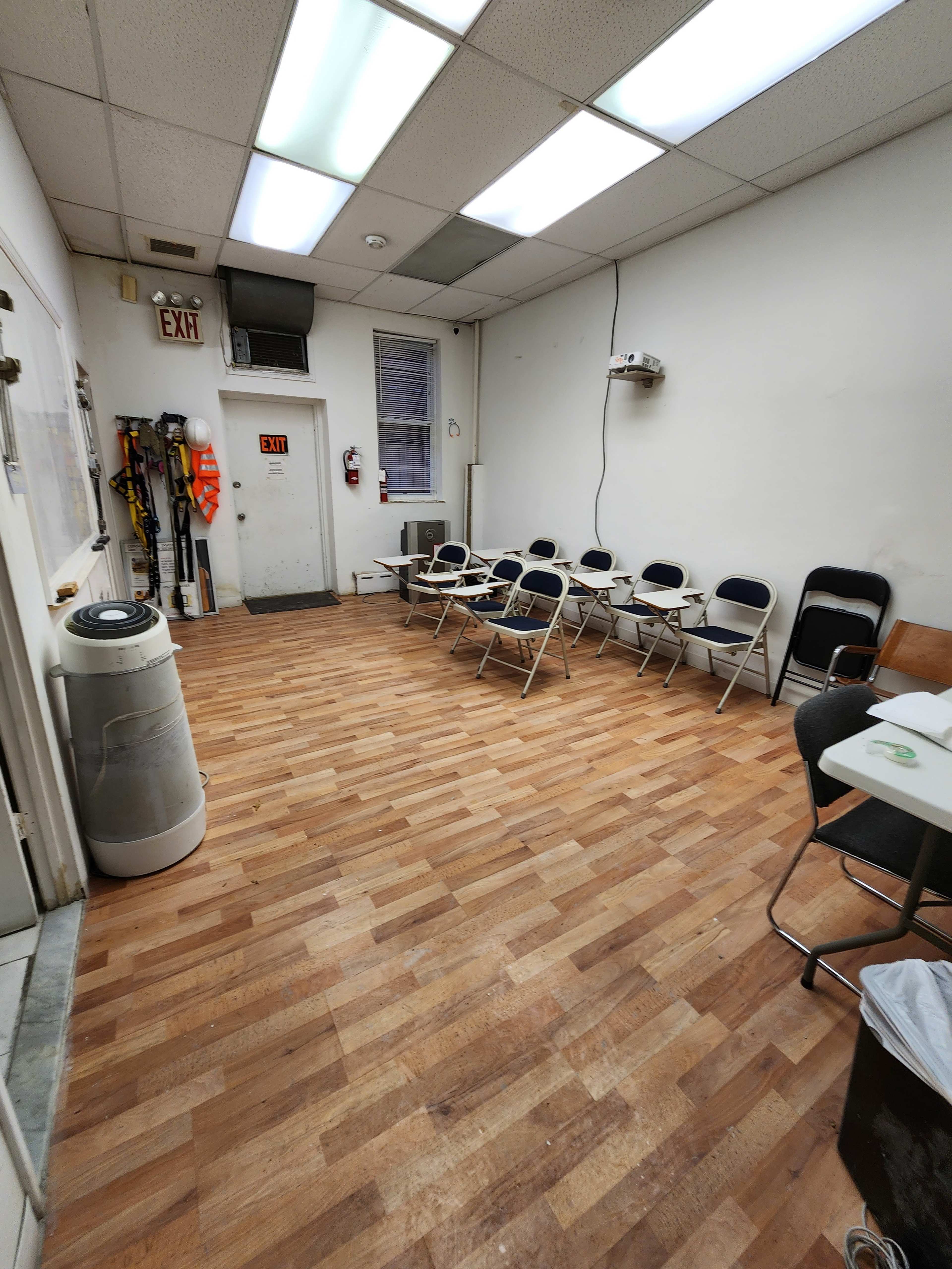An empty room features a wooden floor, a few folding chairs set against the walls, and a door leading outside.