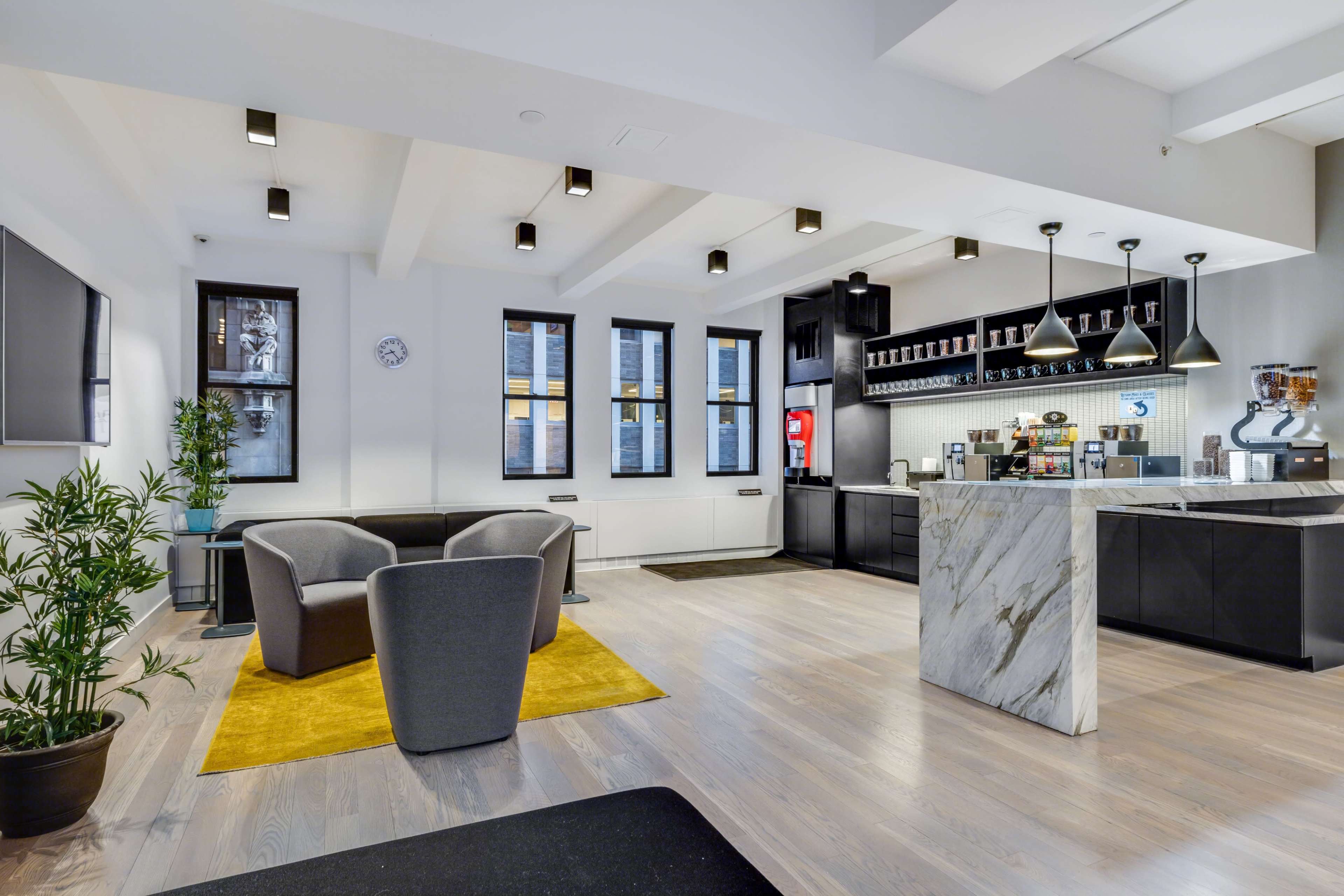 A modern lounge area featuring gray chairs, a yellow rug, and a kitchen countertop with coffee machines and a soda dispenser.