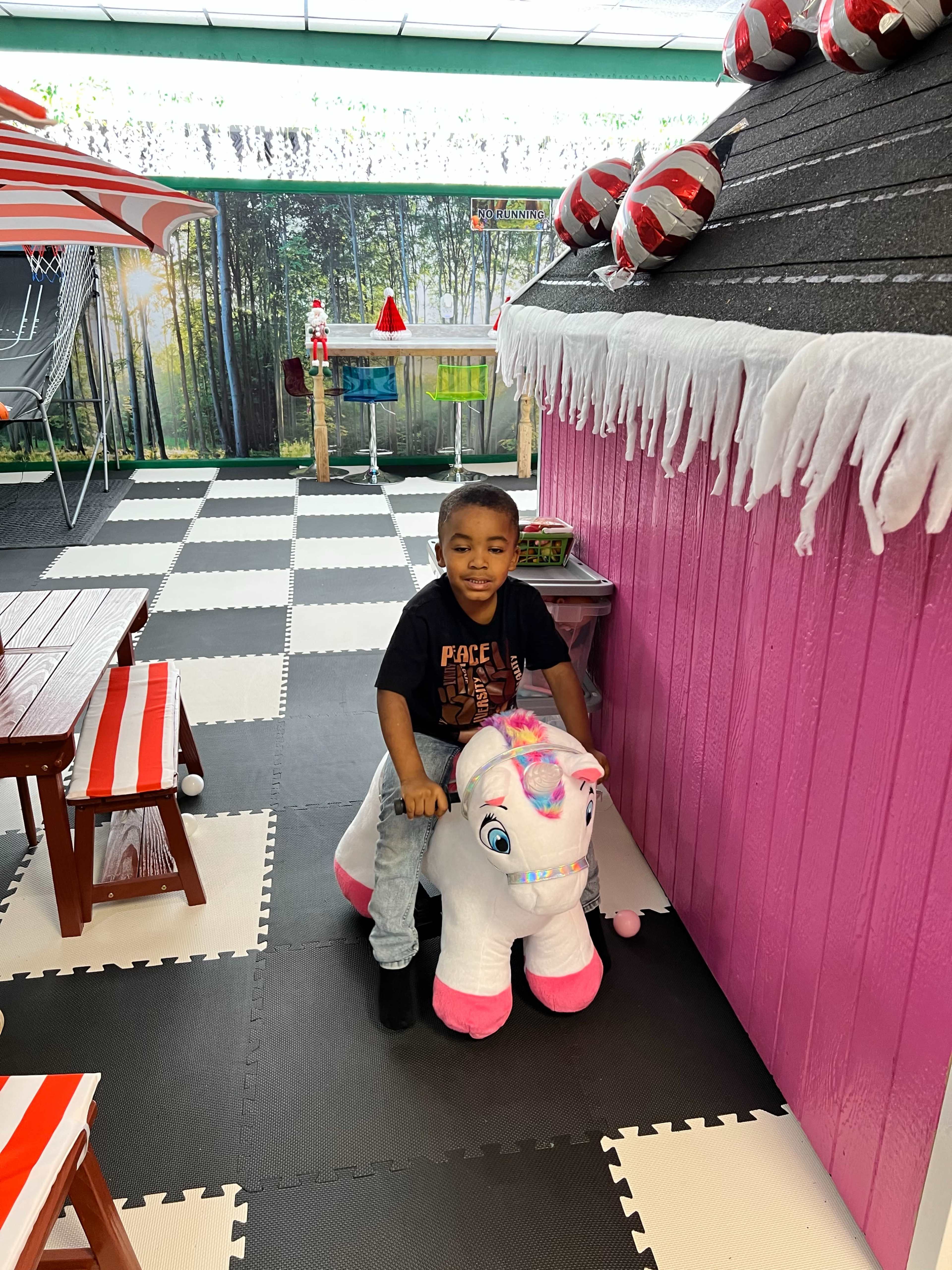 A child sits on a toy unicorn in a colorful play area with a pink playhouse.