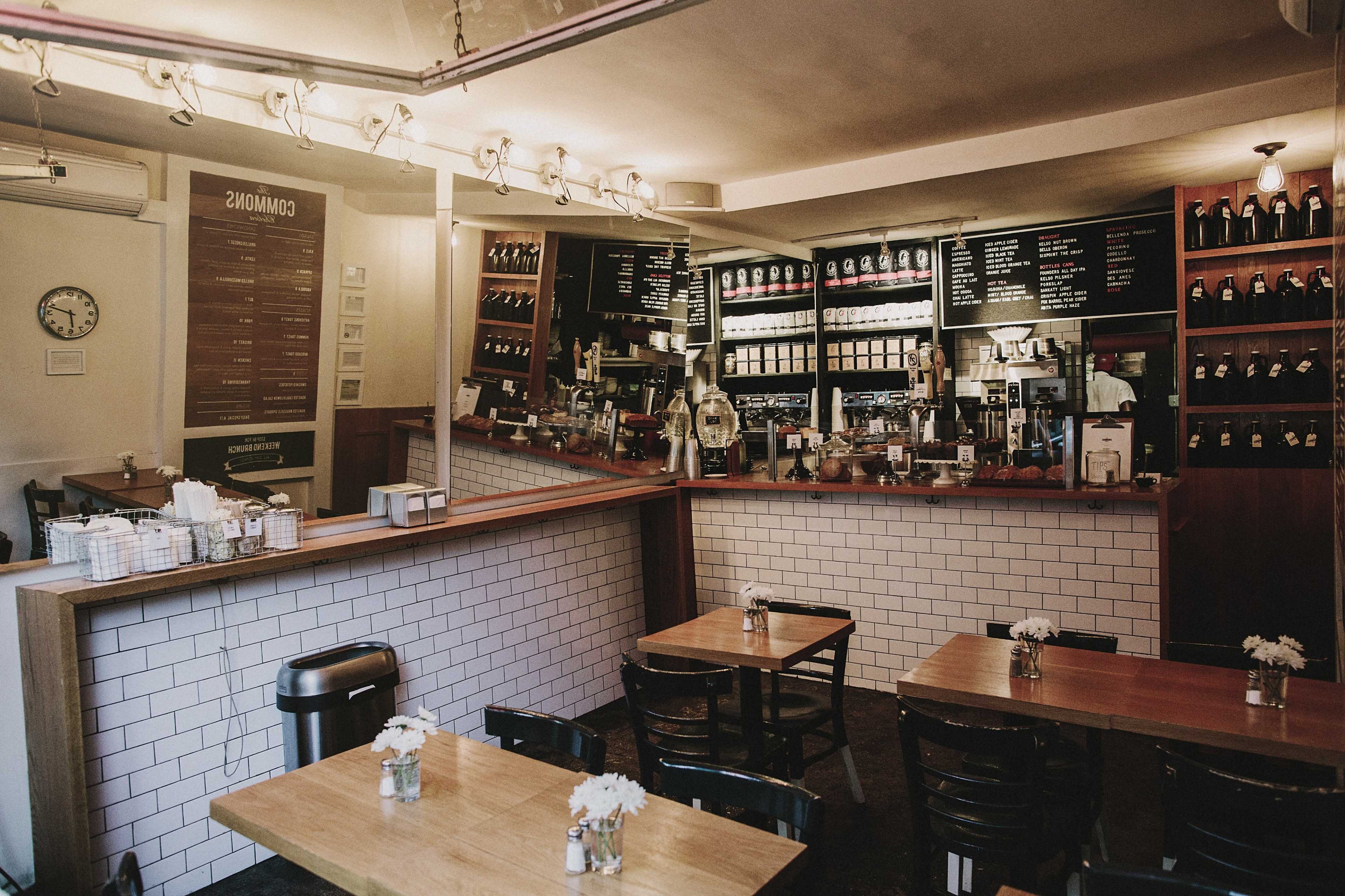 A cozy cafe interior with wooden tables, a barista counter displaying coffee machines, and a wall of shelves filled with various beverages.