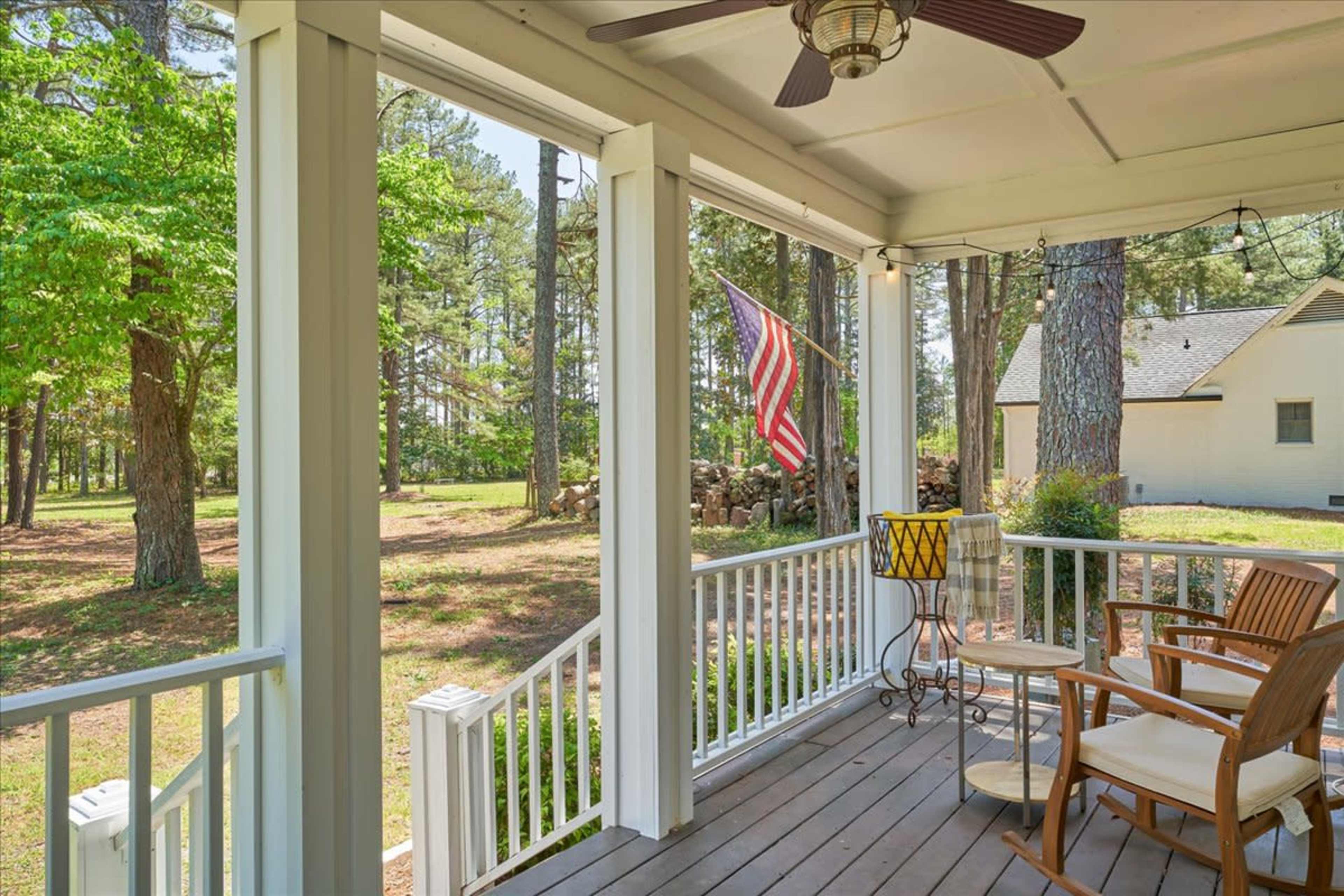 A porch with white railings, a ceiling fan, two chairs, and an American flag waving in the breeze, surrounded by trees and a house in the background.