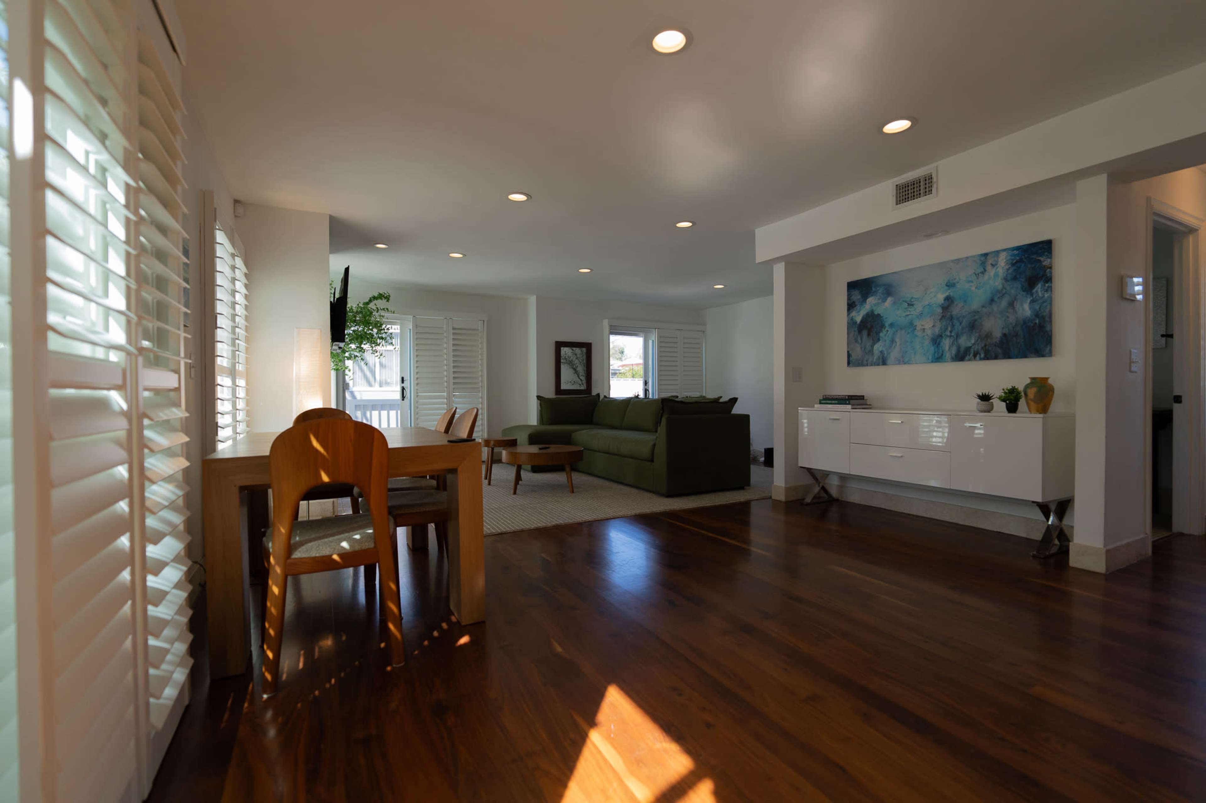 The image shows a bright interior living space featuring a dining table, a green sofa, and a large window with plantation shutters.