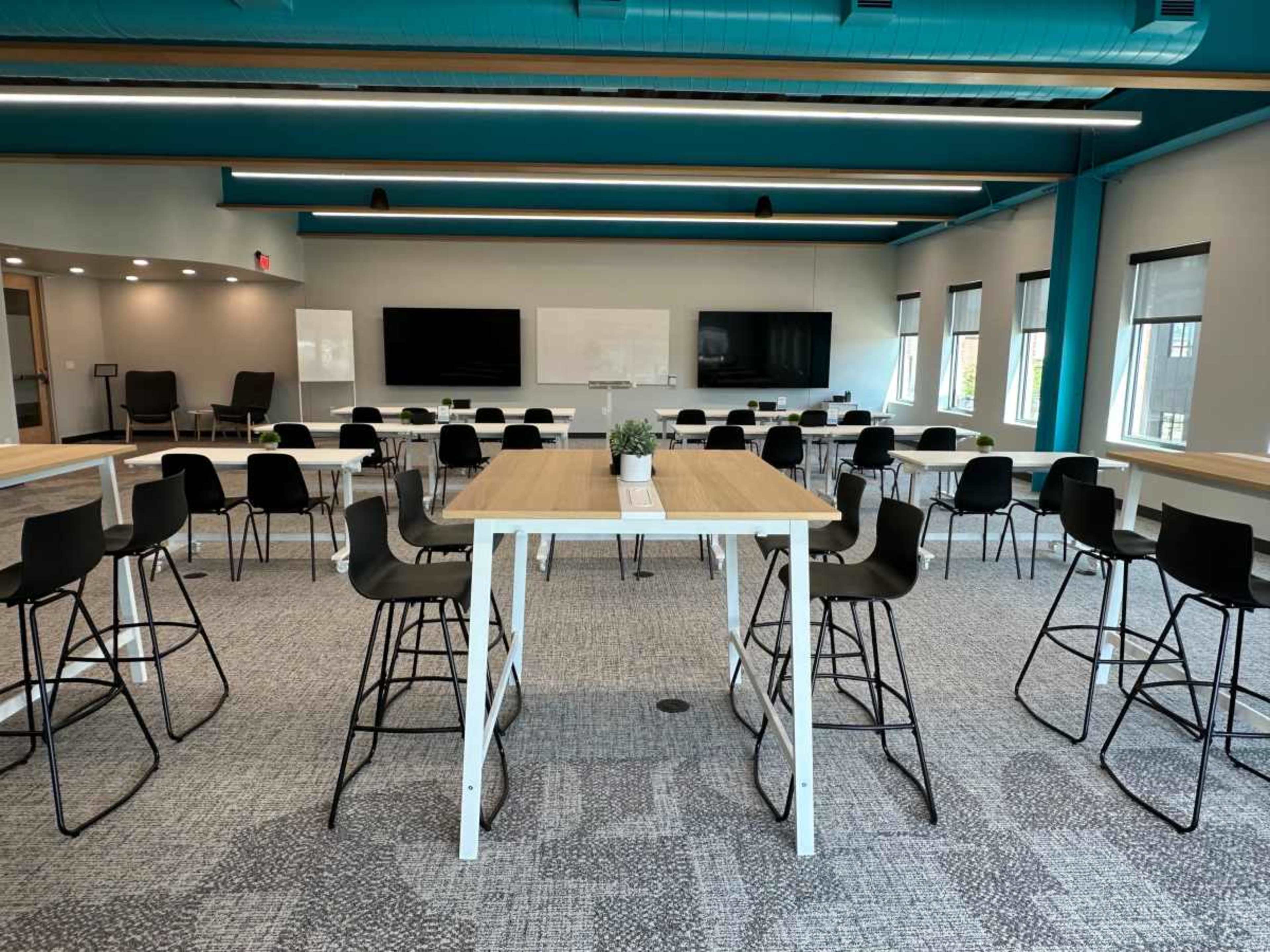 A modern conference room features several long tables surrounded by high-backed chairs, with large screens and windows along the walls.