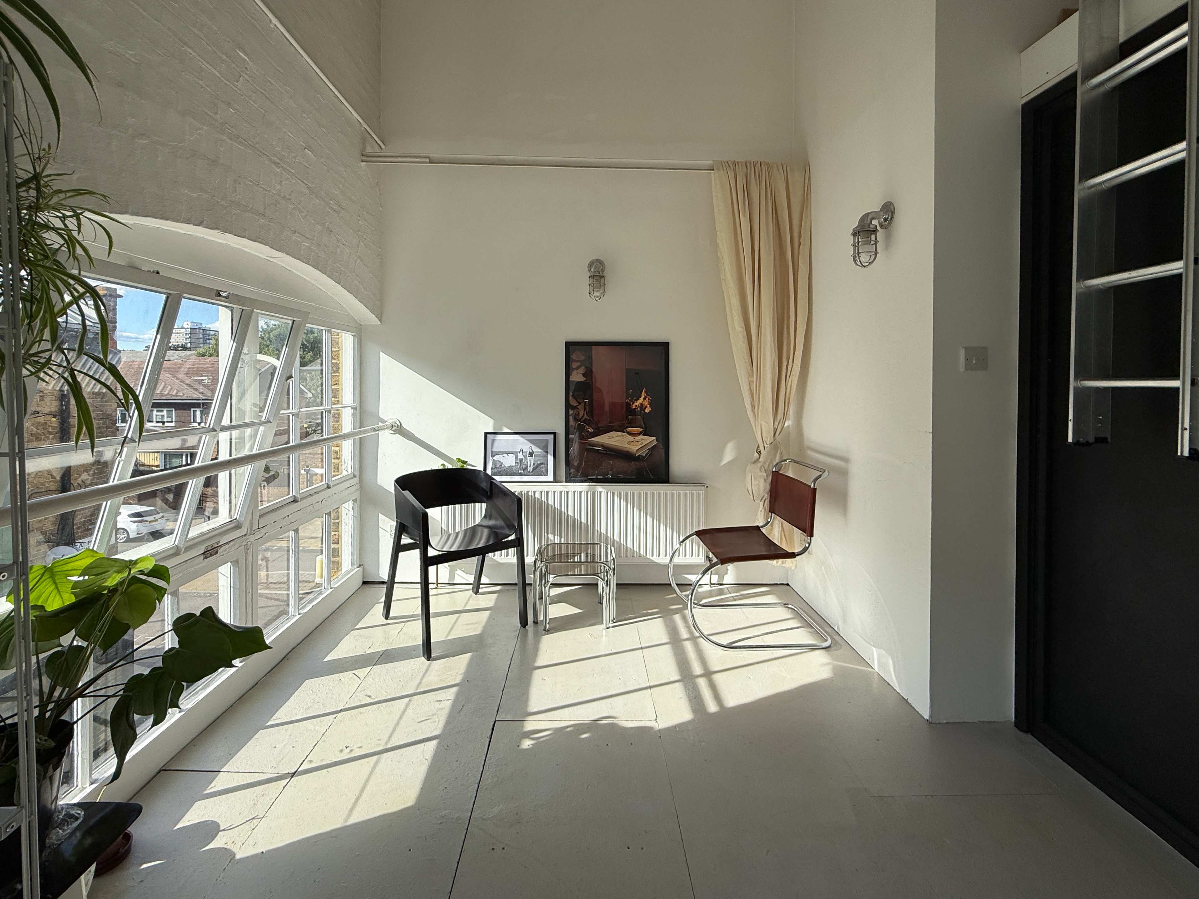 A bright room features a white wall, large windows, a table, and two chairs, along with a potted plant and artwork on display.