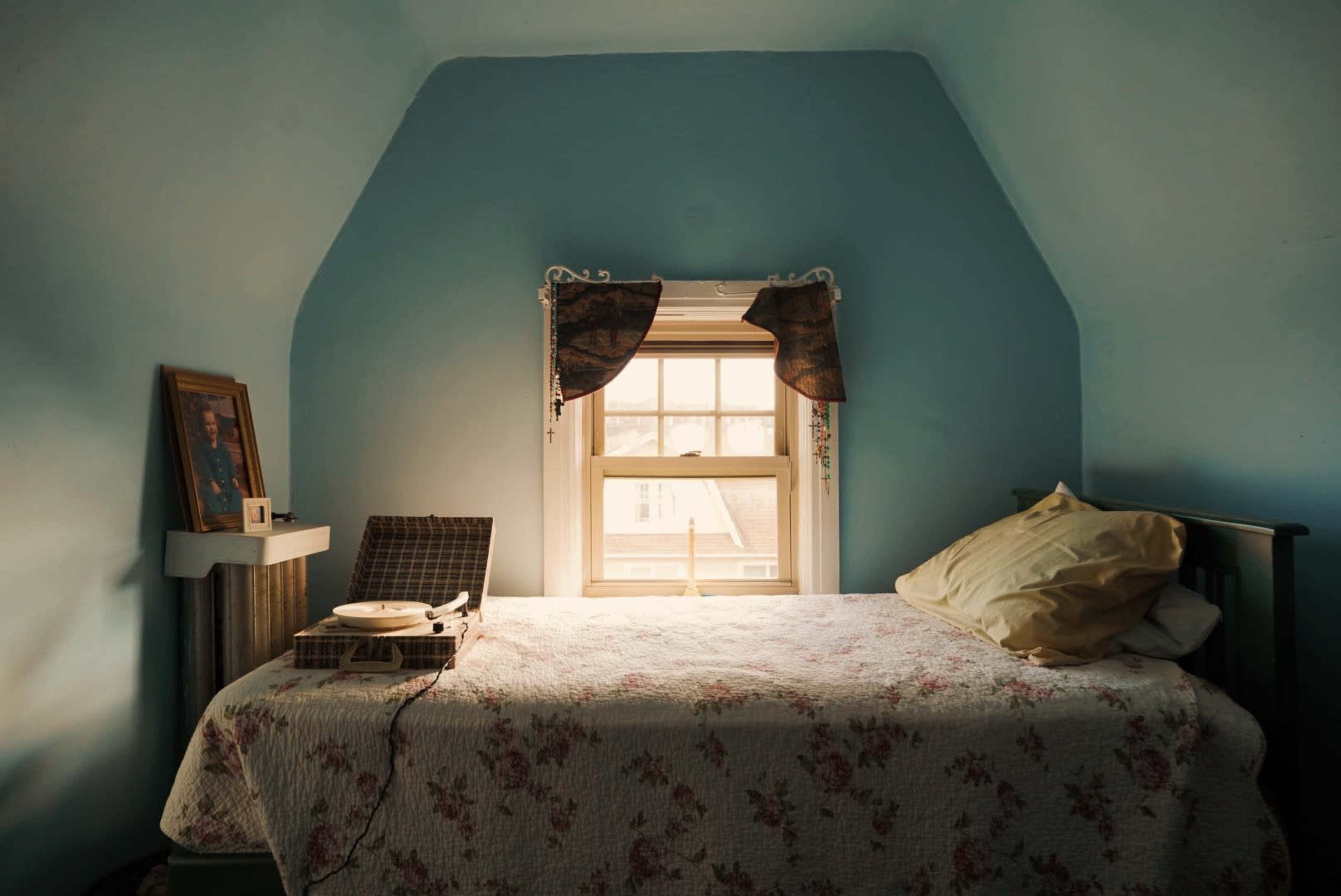 A small bedroom features a bed with floral bedding, a record player beside it, and a window with light filtering through curtained drapes.