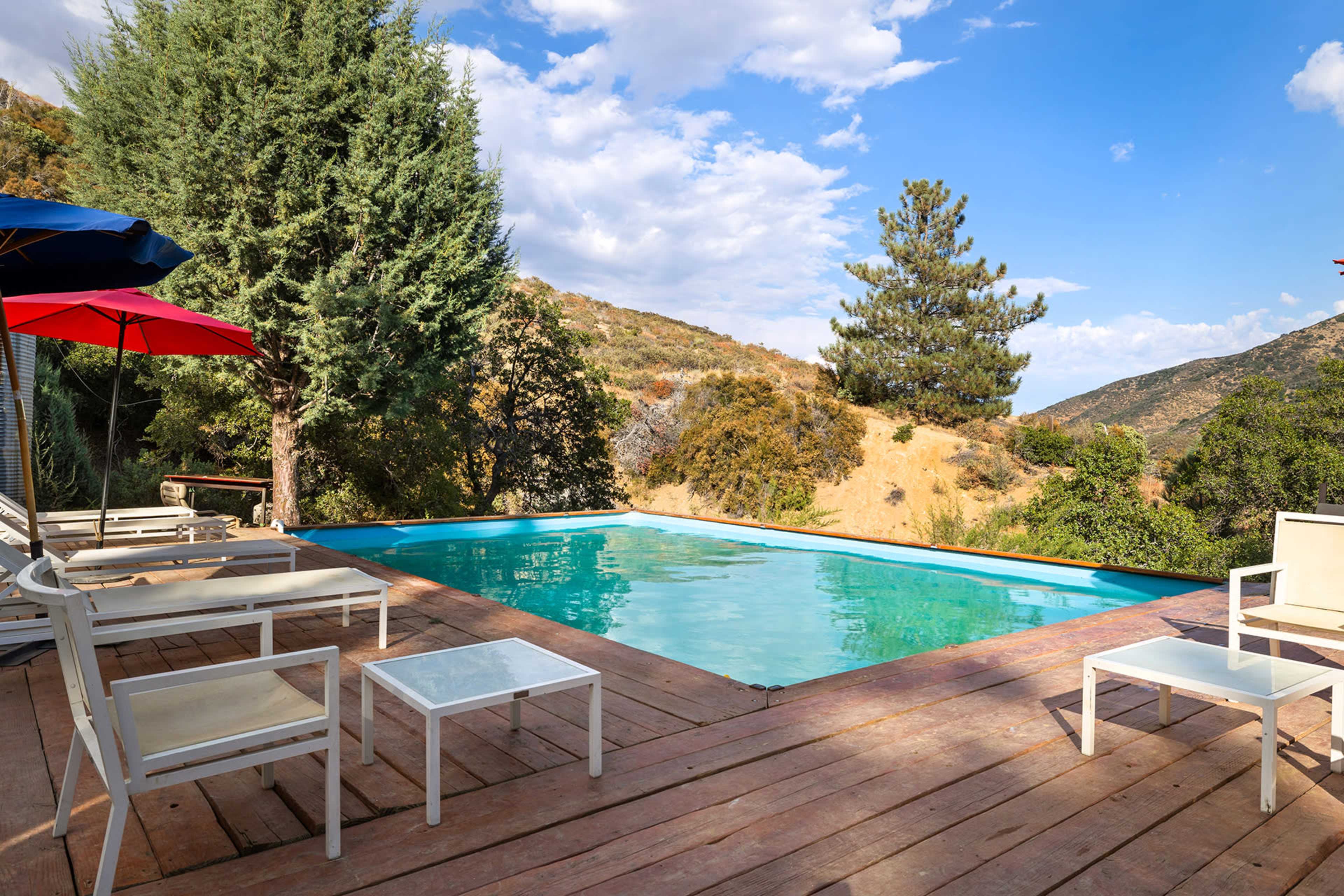 A swimming pool is situated on a wooden deck surrounded by trees and hills under a blue sky with scattered clouds.