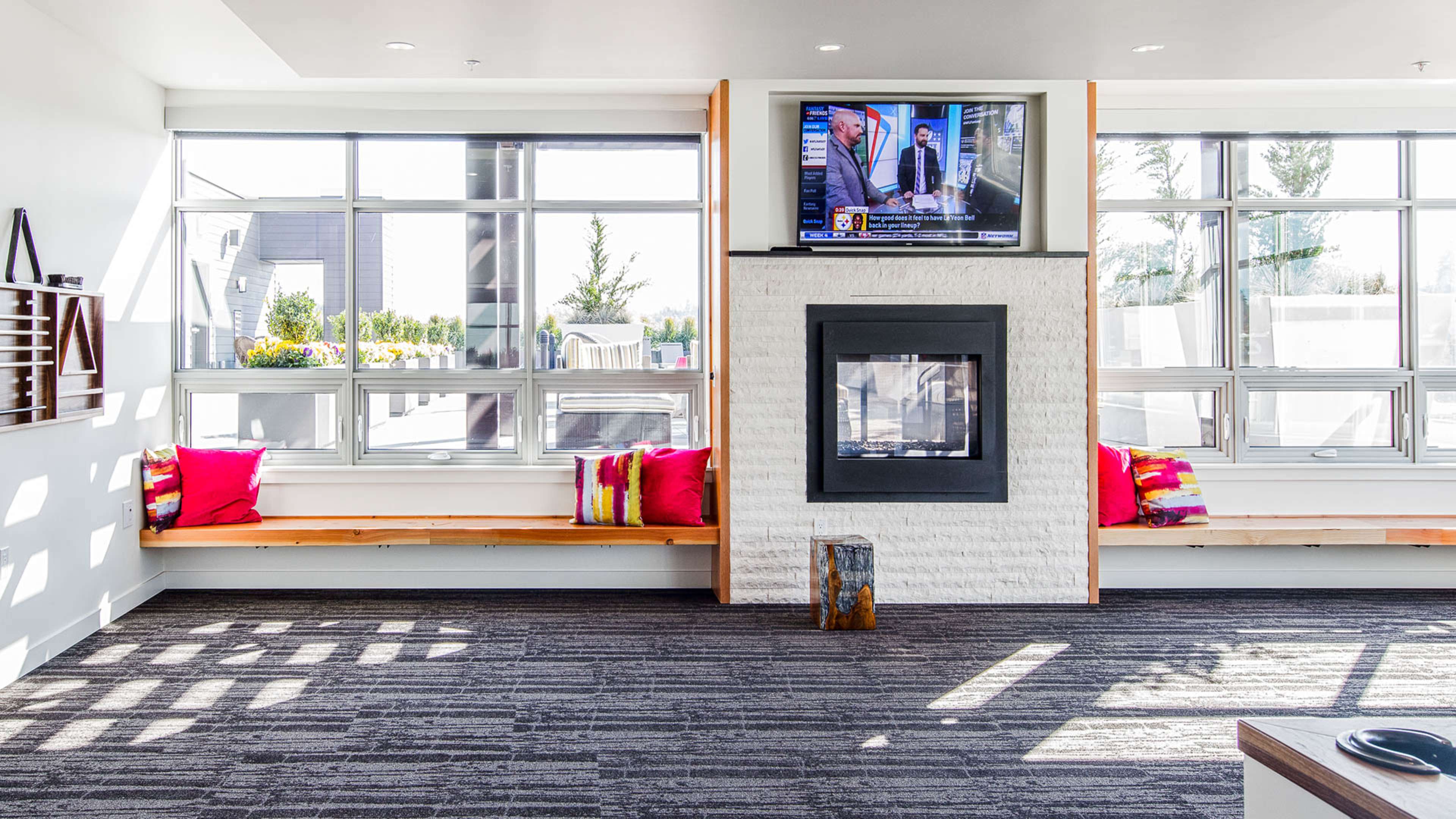 A modern interior room with a television mounted above a fireplace, featuring two benches with colorful cushions along the windows and a decorative wooden stool.