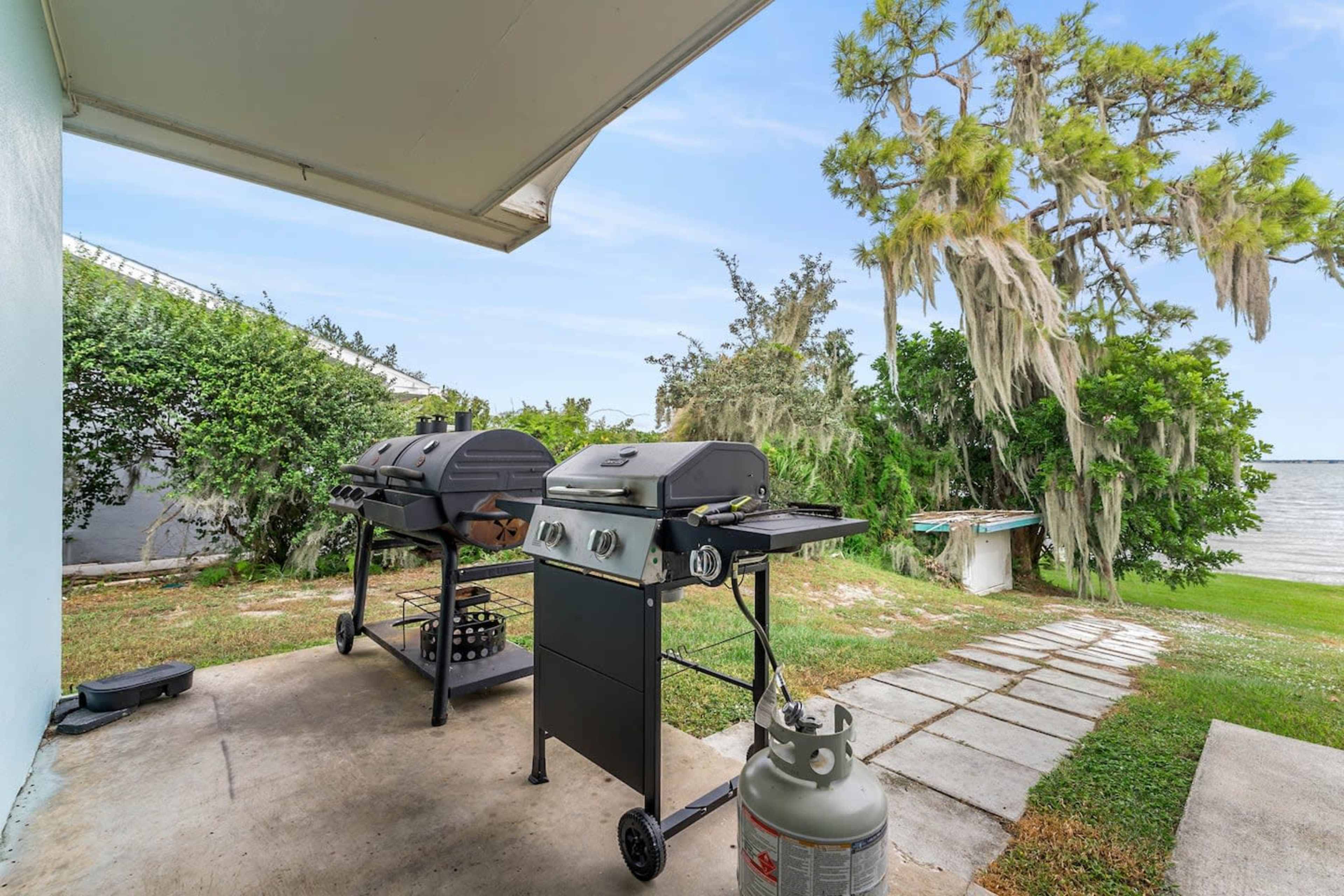 Two gas grills are set up on a concrete patio, overlooking a body of water with trees in the background.