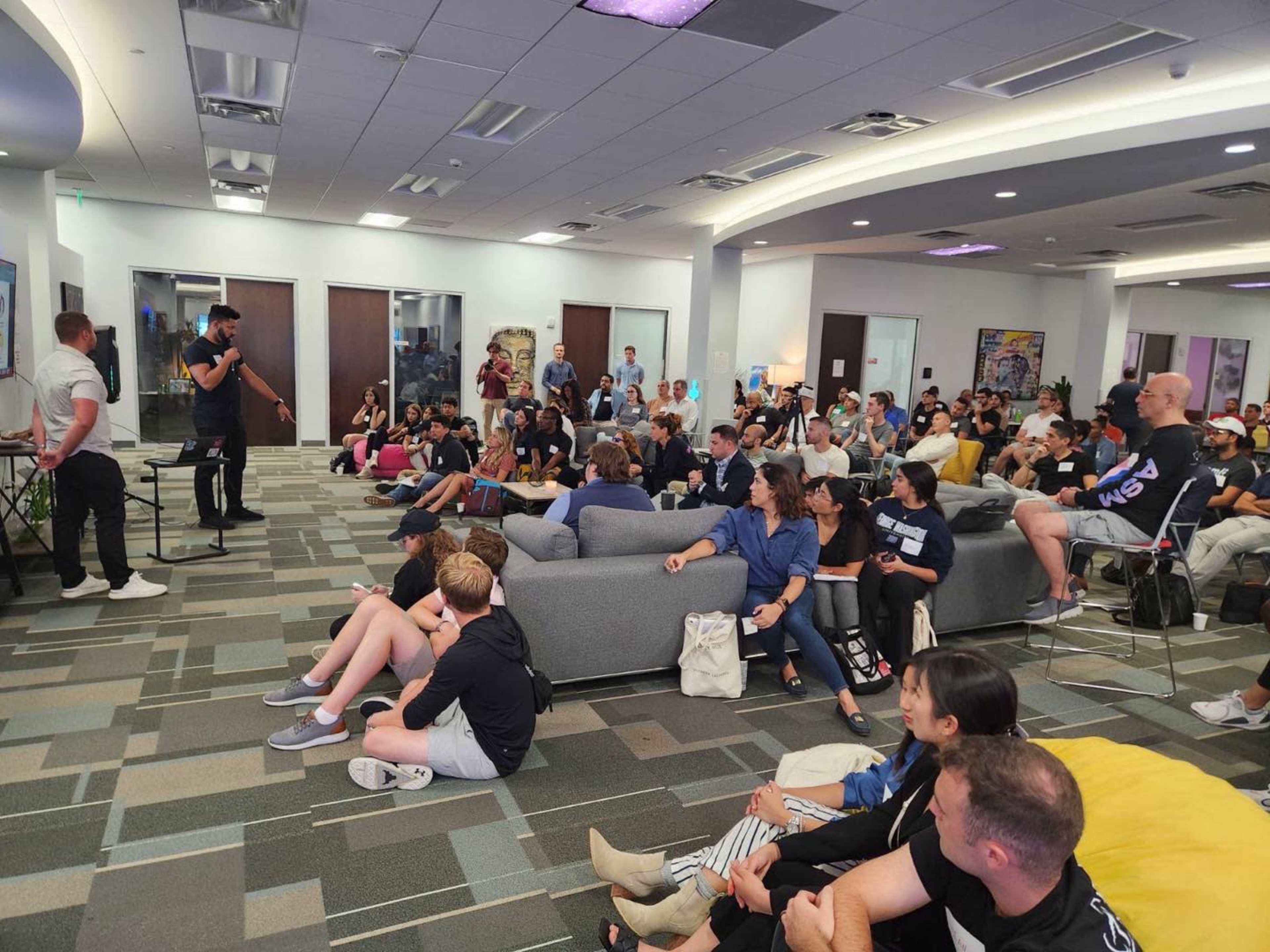 A large group of people listens to a speaker in a modern office space with gray sofas and colorful bean bags.