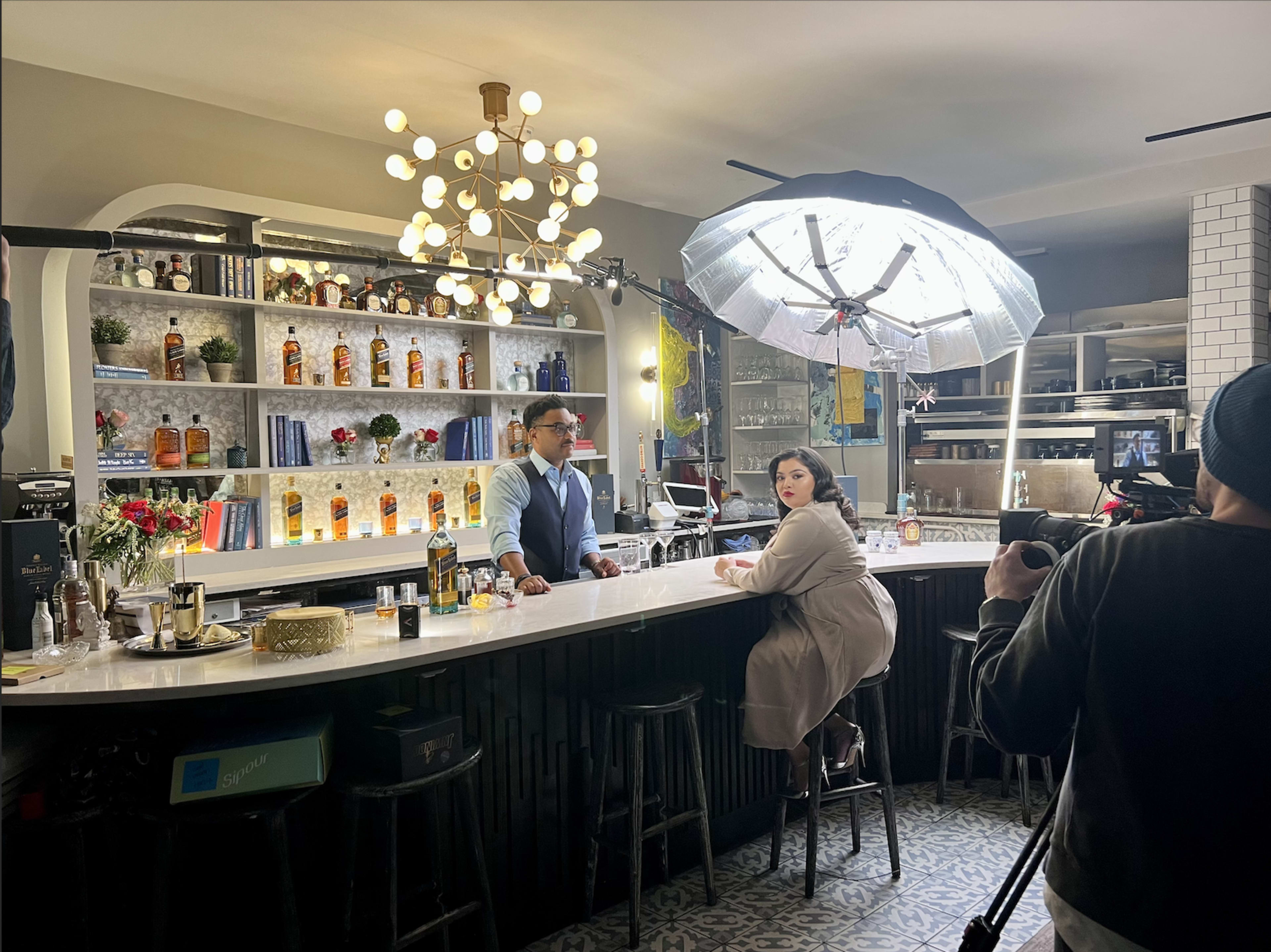 A bartender and a woman sit at a well-lit bar surrounded by shelves of various spirits and decorations, with a large softbox light directing focus on them.
