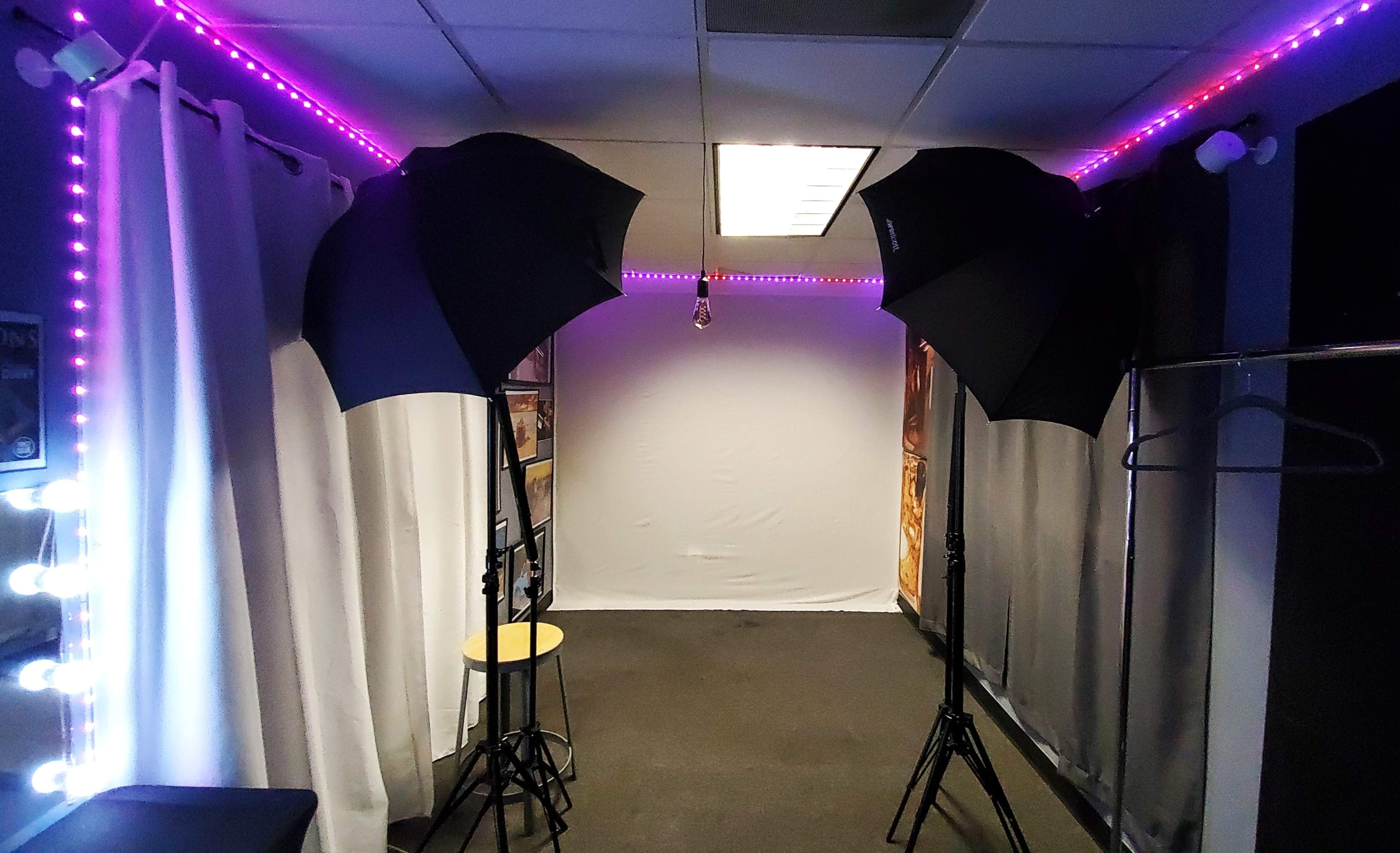 The image shows a brightly lit photography studio with two softbox lights, a plain backdrop, and a stool positioned in front of it.
