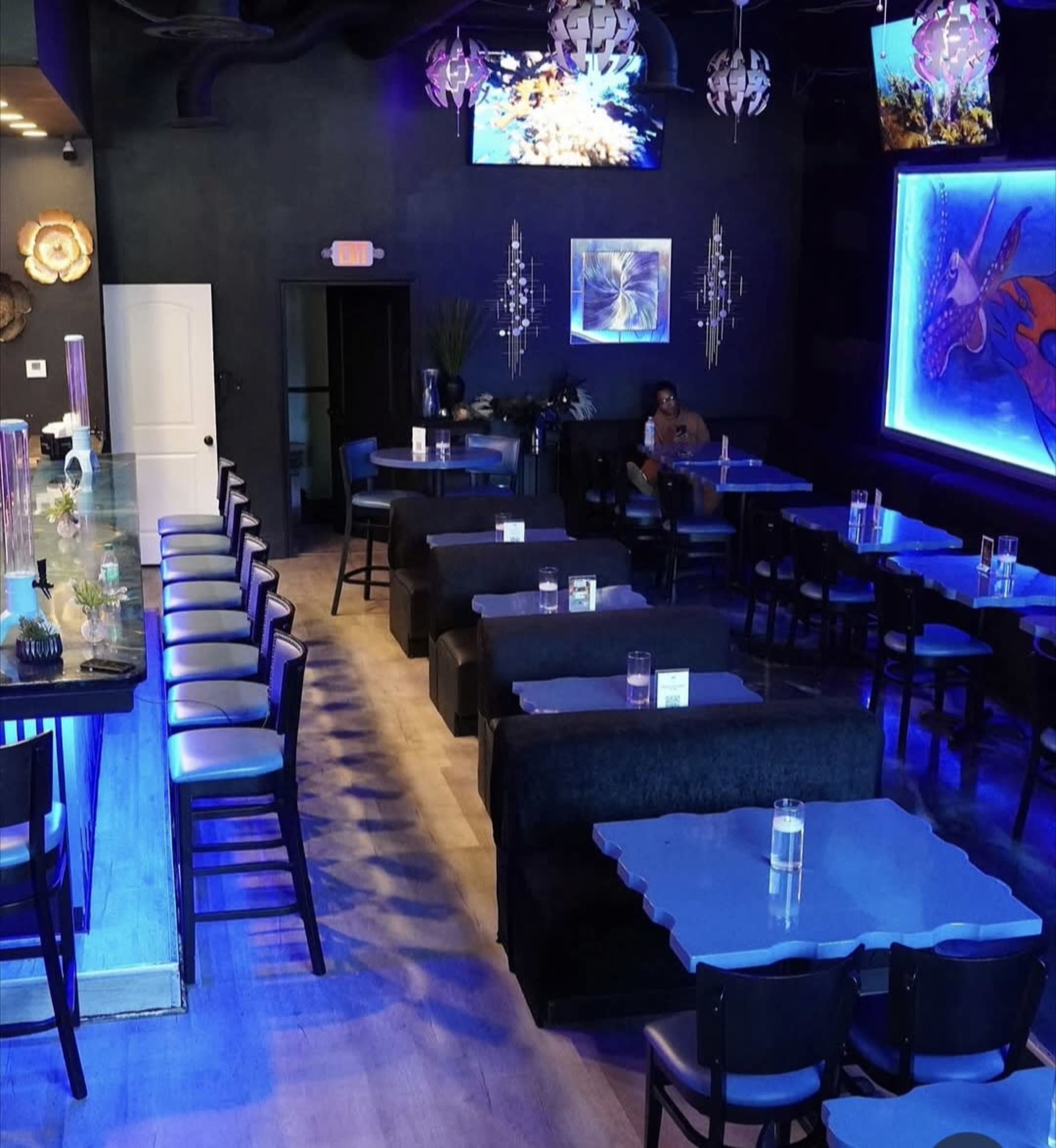The image shows the interior of a modern restaurant with dark walls, blue lighting, and seating arranged at booths and a bar.