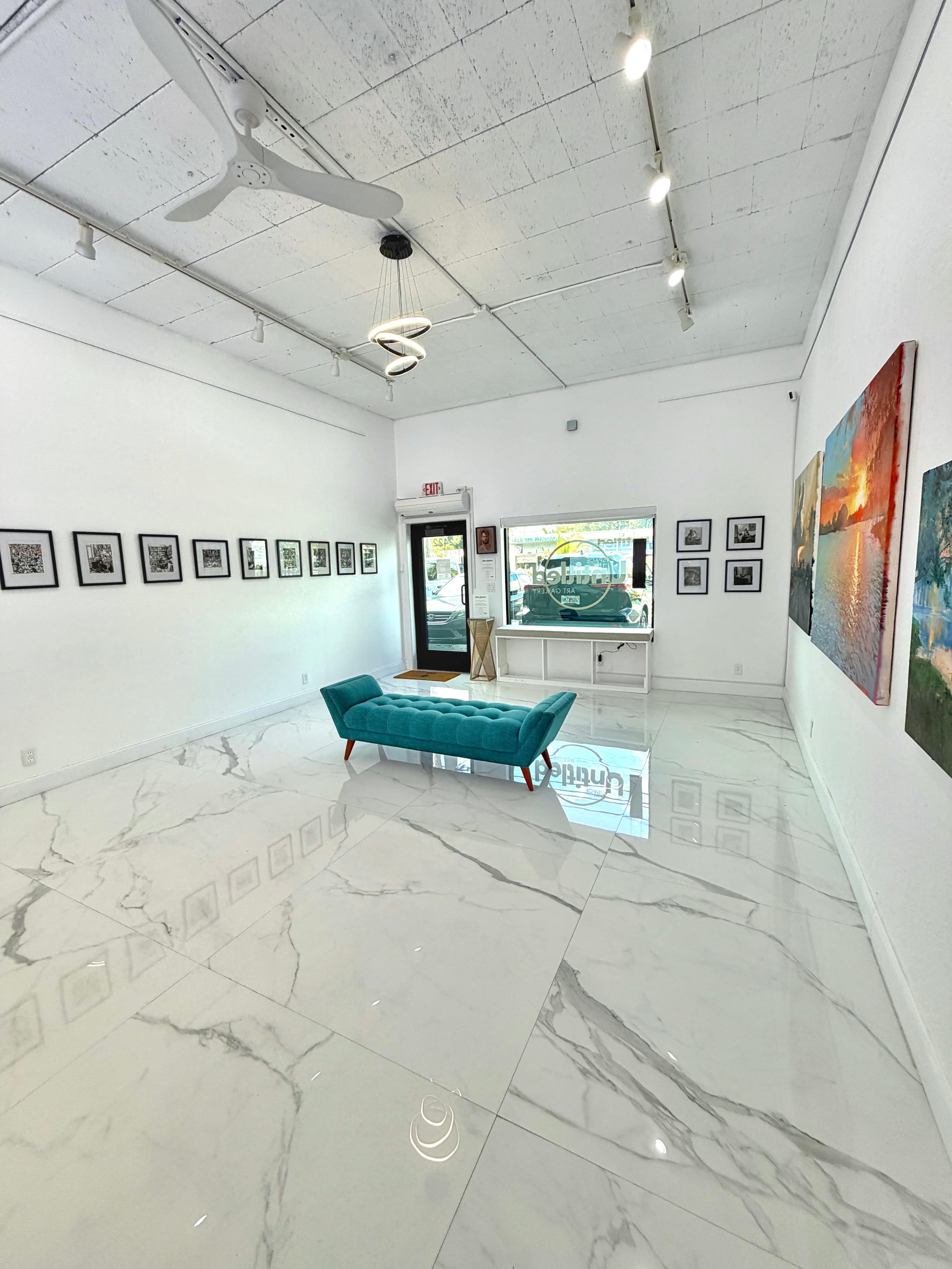 The image shows a modern art gallery featuring a white interior with marble flooring, displaying framed photographs on the walls and a turquoise couch in the center.