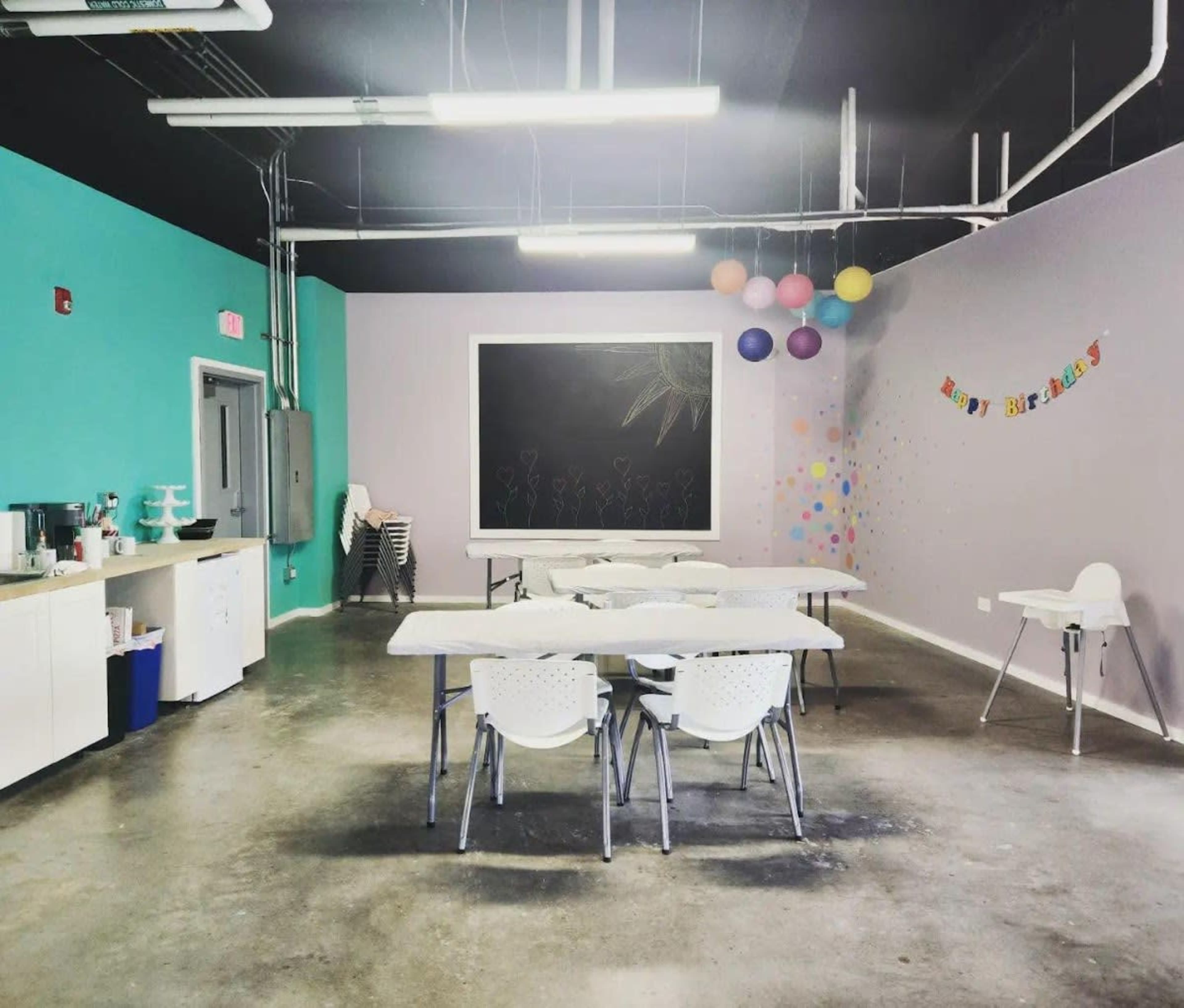 A spacious room with multiple tables and chairs arranged, a chalkboard on the wall, and colorful decorations including balloons and a birthday banner.