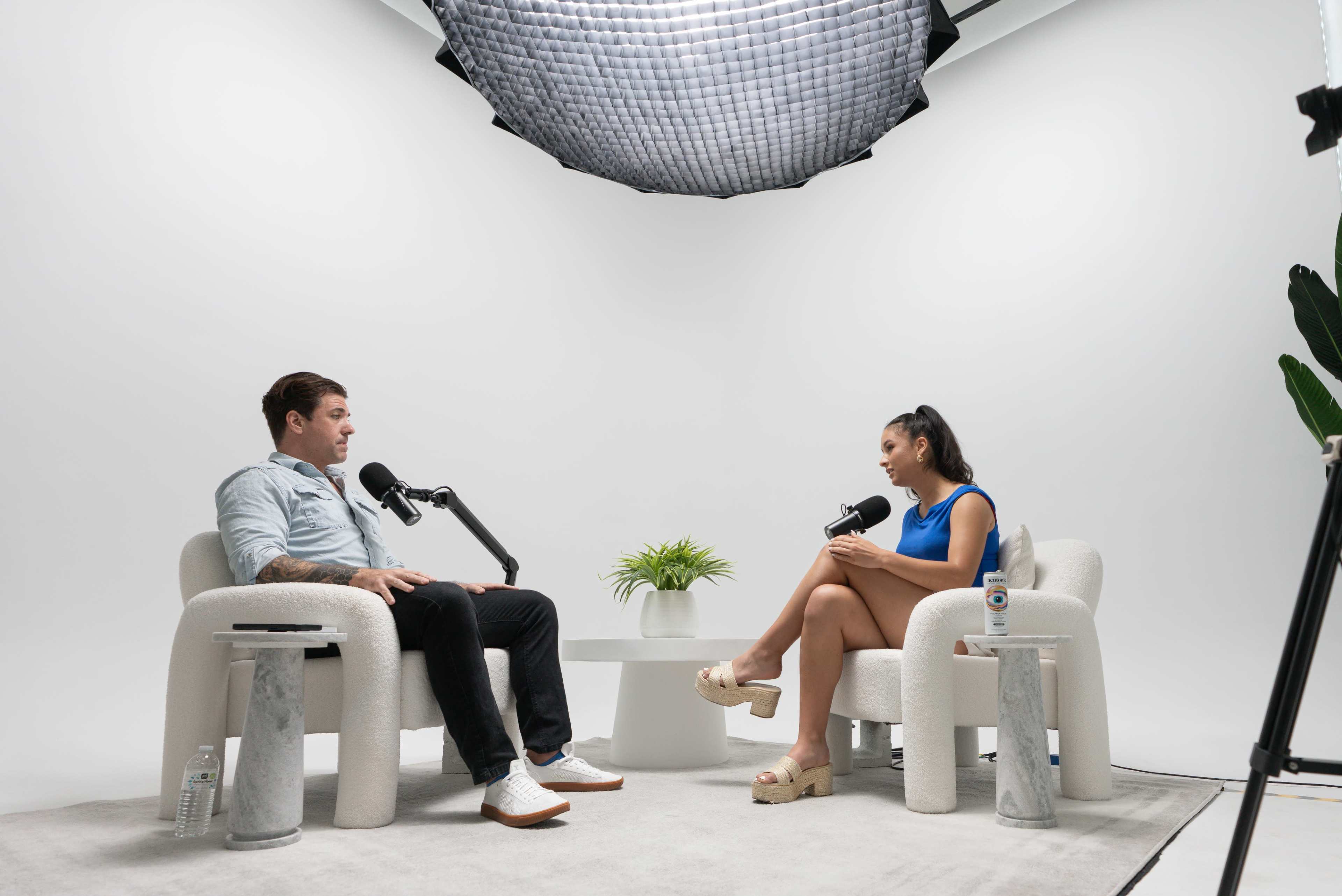 Two people are seated in minimalist chairs, engaged in a conversation, with microphones positioned between them in a well-lit studio setting.