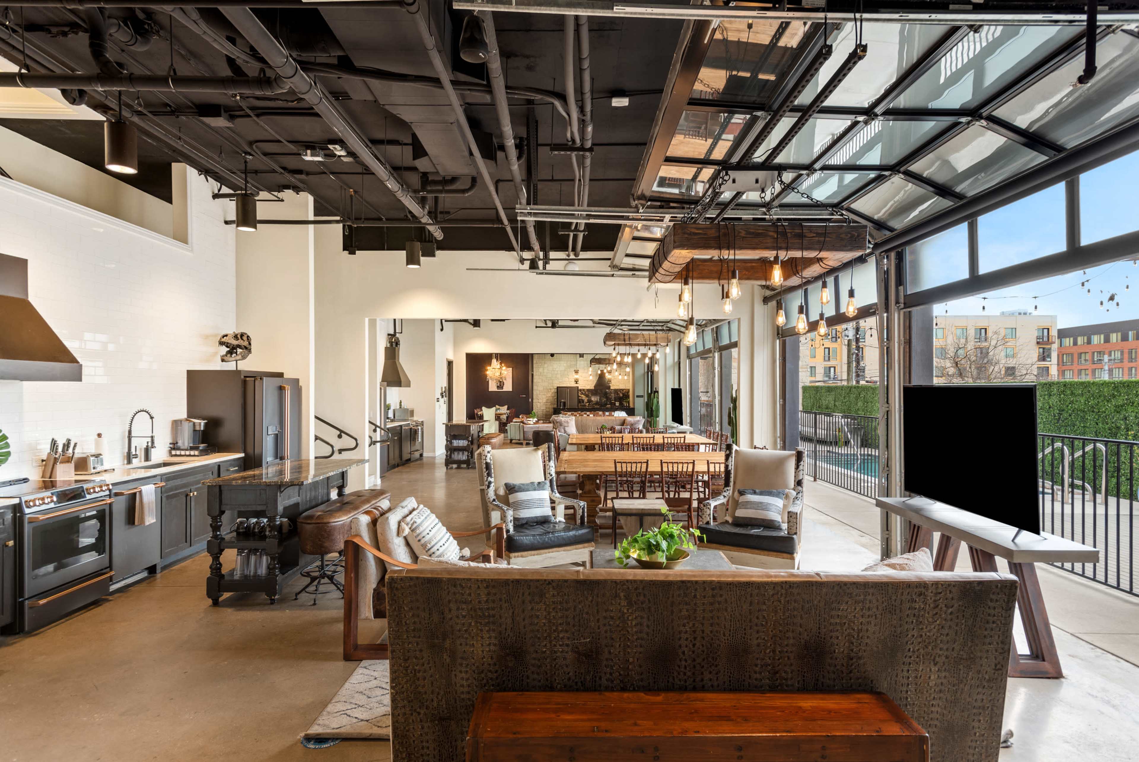 The image shows a modern, open-concept living space with a kitchen, dining area, and lounge section featuring large windows and an industrial aesthetic.