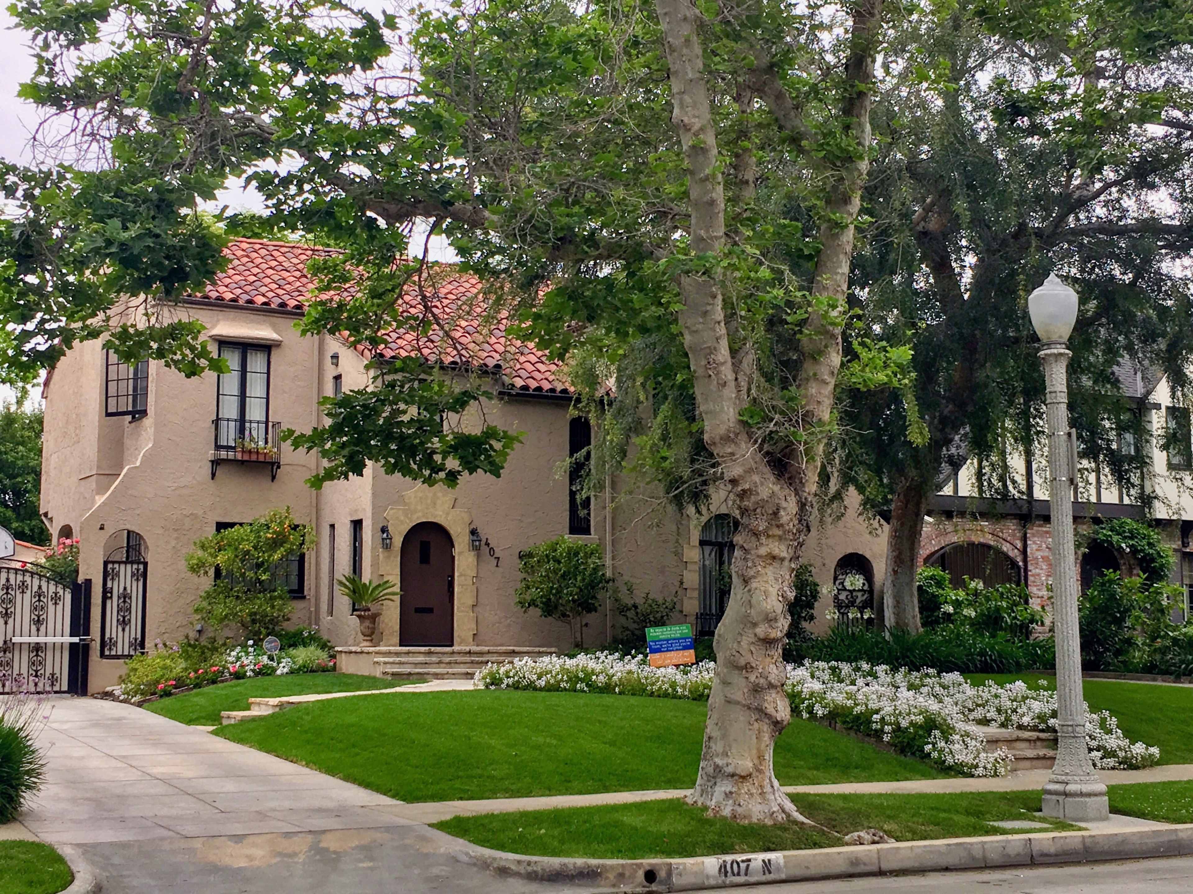 The image shows a two-story stucco house with a red tiled roof, surrounded by trees and a landscaped yard featuring white flowers.