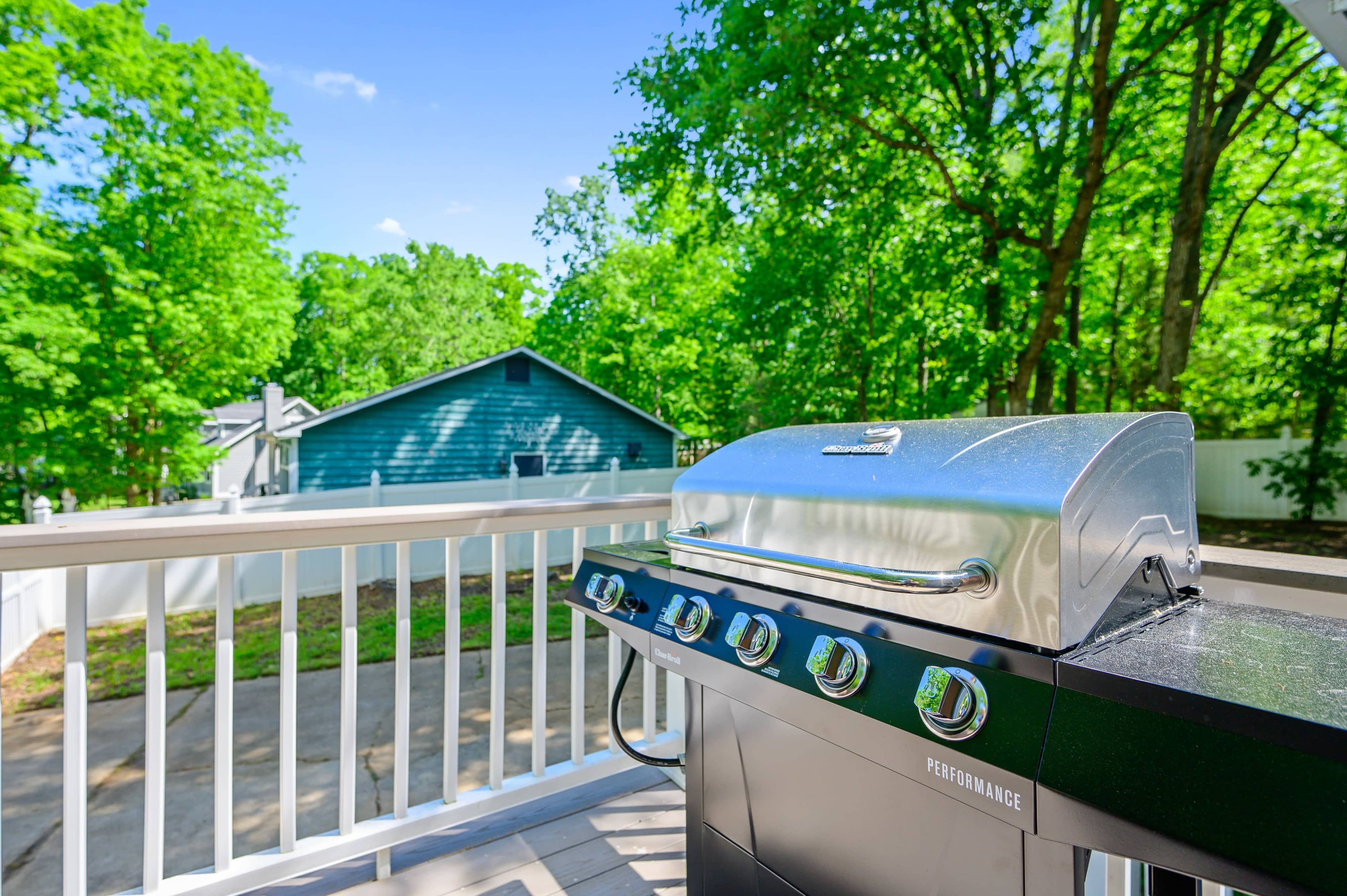 A stainless steel grill is positioned on a deck overlooking green trees and a blue house in the background.