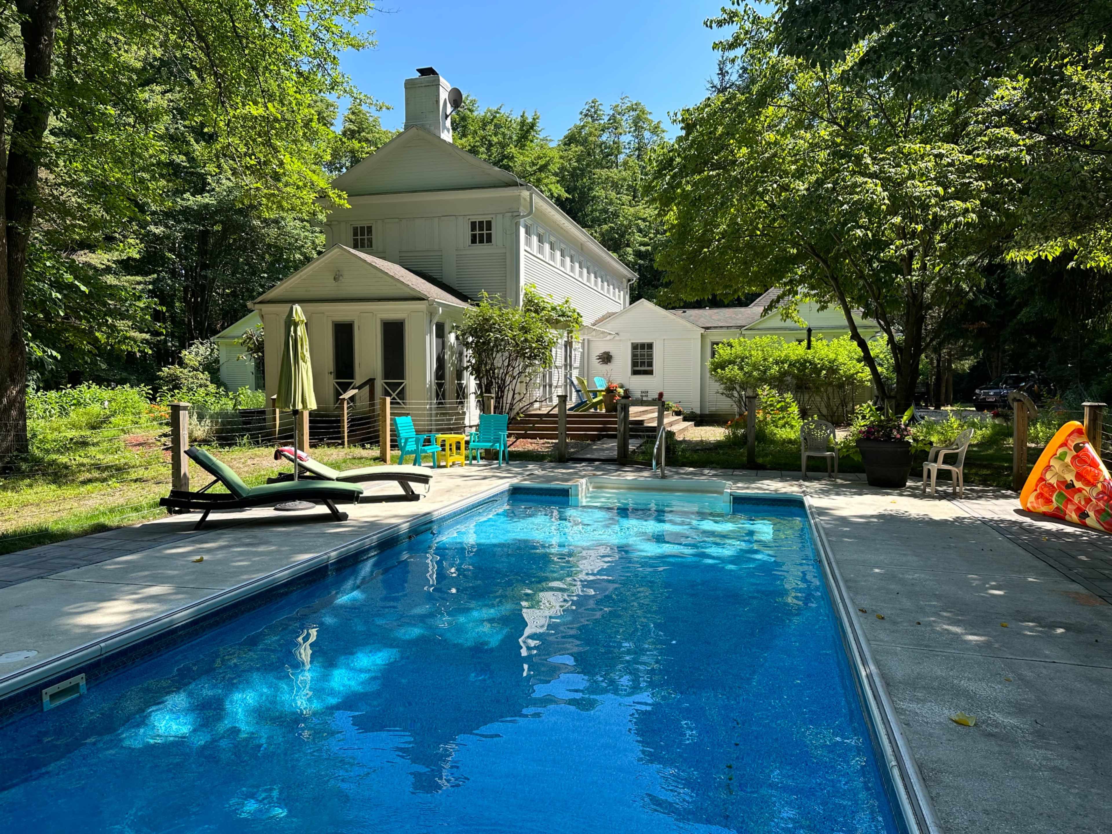 A clear blue swimming pool is situated in the foreground, with a white house and green trees in the background.