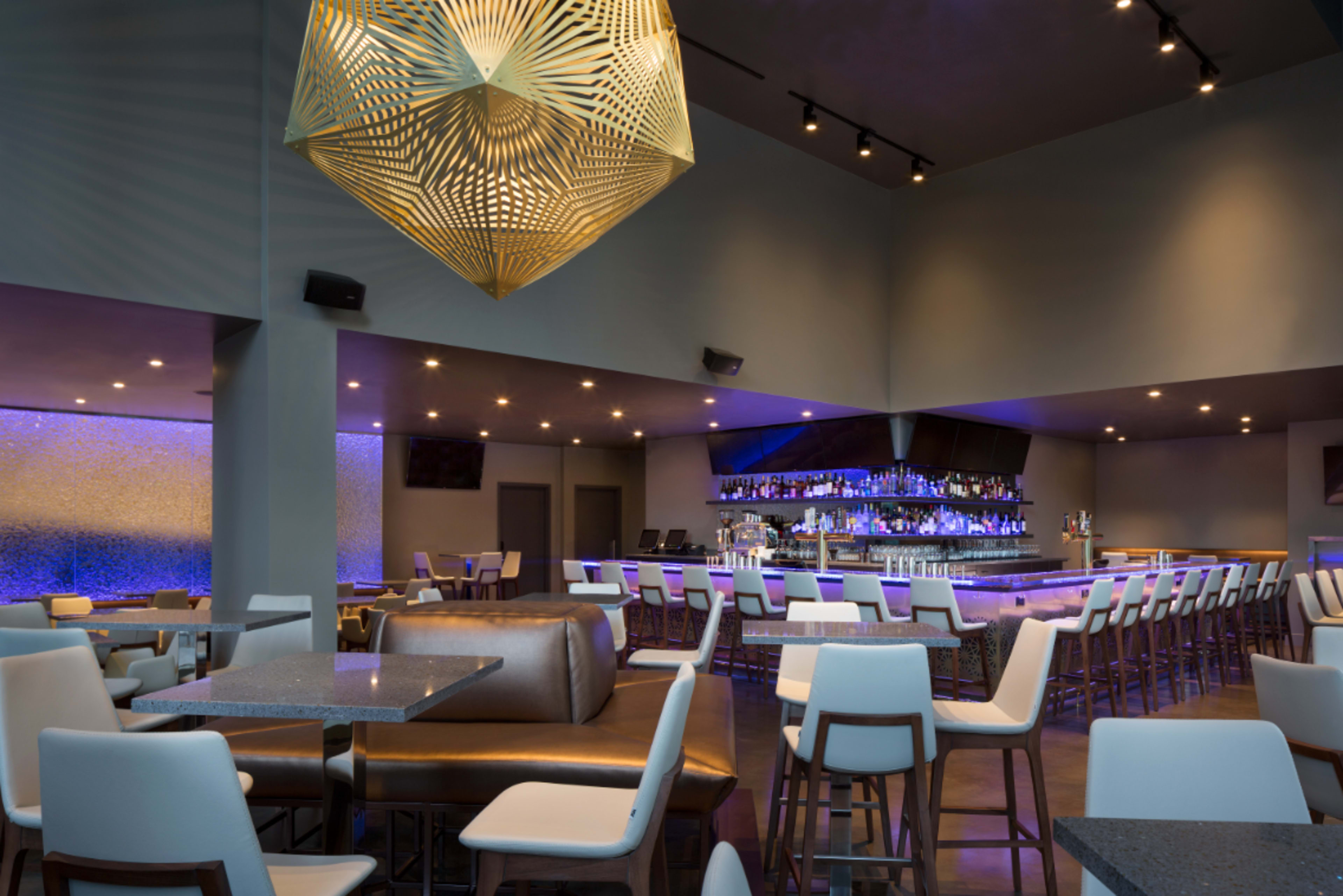 A modern bar and dining area with a stylish geometric light fixture overhead, a long bar stocked with drinks, and multiple seating arrangements.