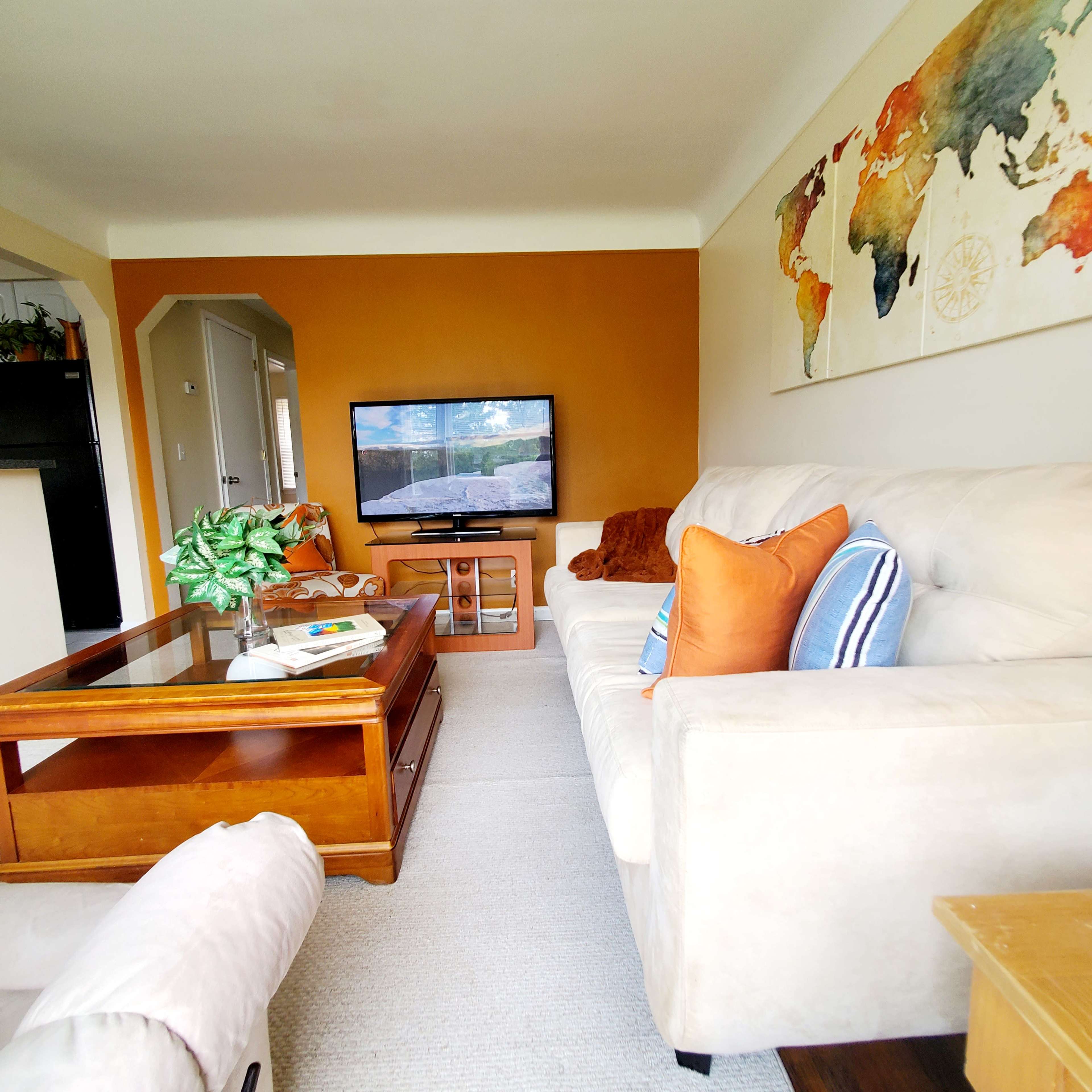 The living room includes a beige sofa with orange and blue accent pillows, a wooden coffee table, and a television against an orange wall.