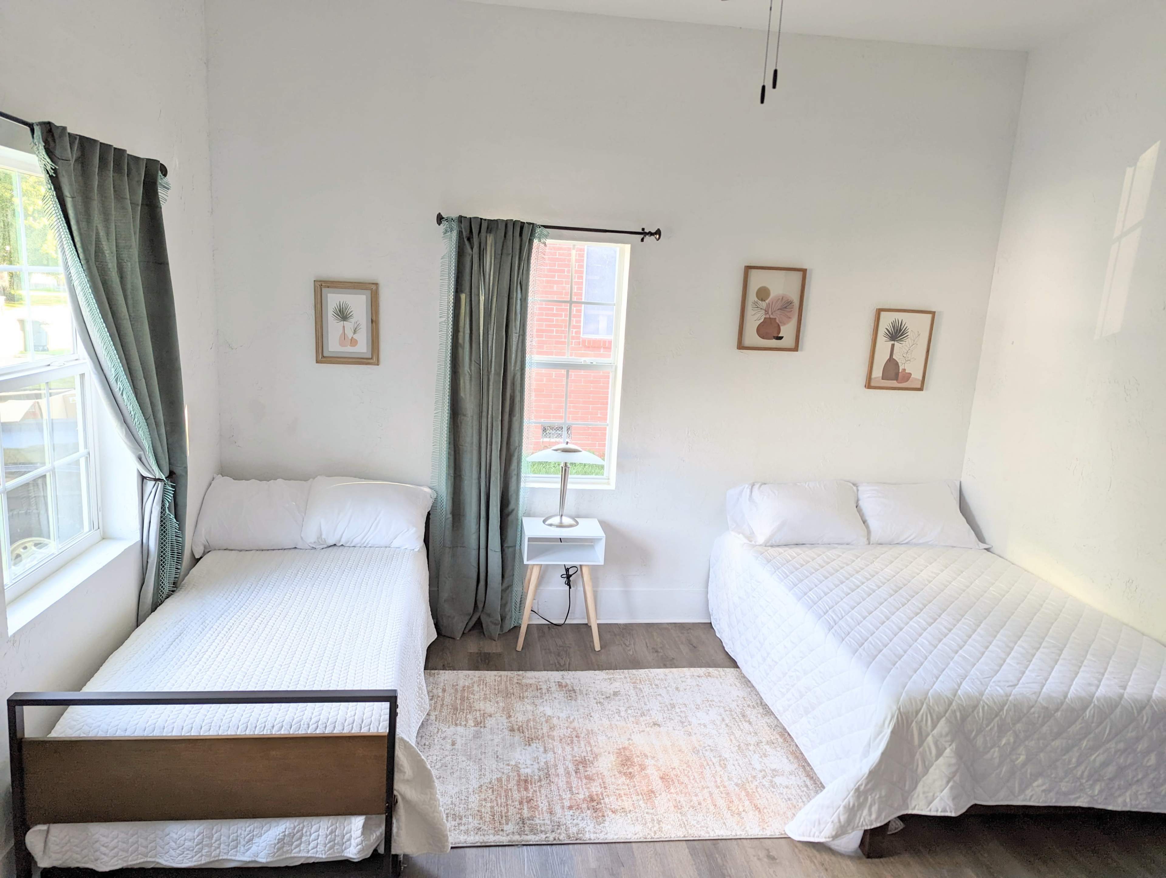 A bright, tidy bedroom featuring two beds, a small bedside table with a lamp, and light green curtains framing the windows.