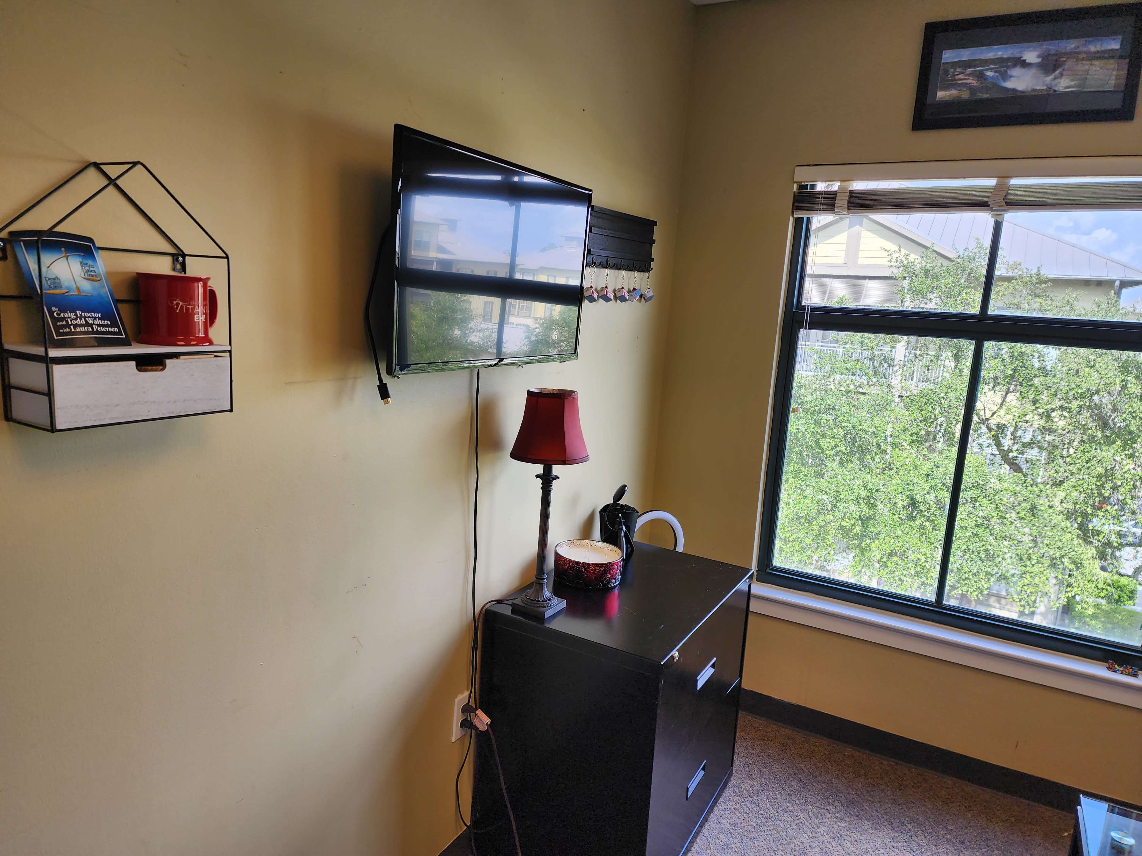 The image shows a small room with a wall-mounted TV, a lamp on a cabinet, a bookshelf with a coffee mug, and a window overlooking greenery.