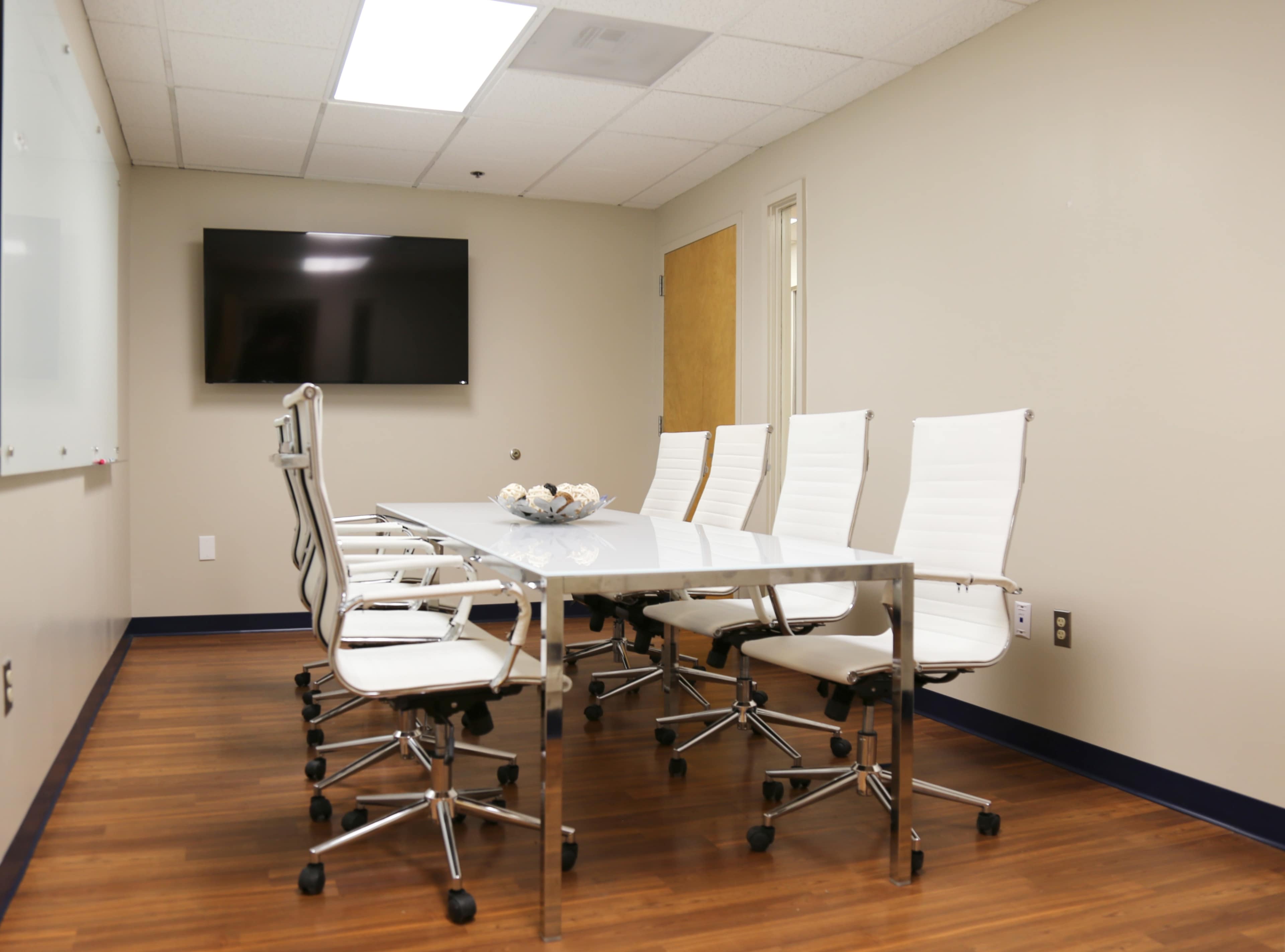 A modern conference room with a glass table and white rolling chairs, featuring a wall-mounted TV and a light-colored wall.
