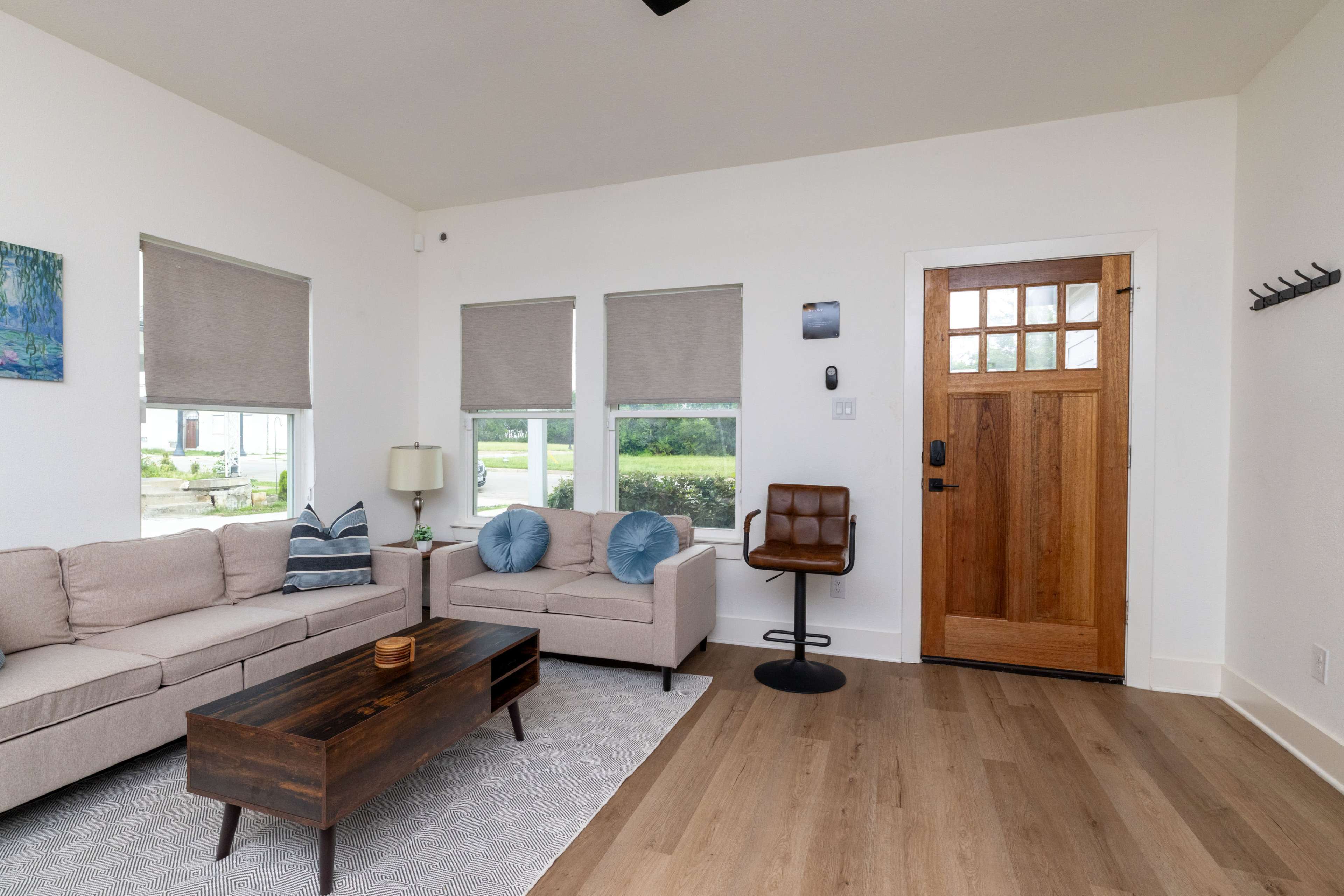 The room features a light-colored sofa set, a wooden coffee table, an entry door, and several windows with shades.