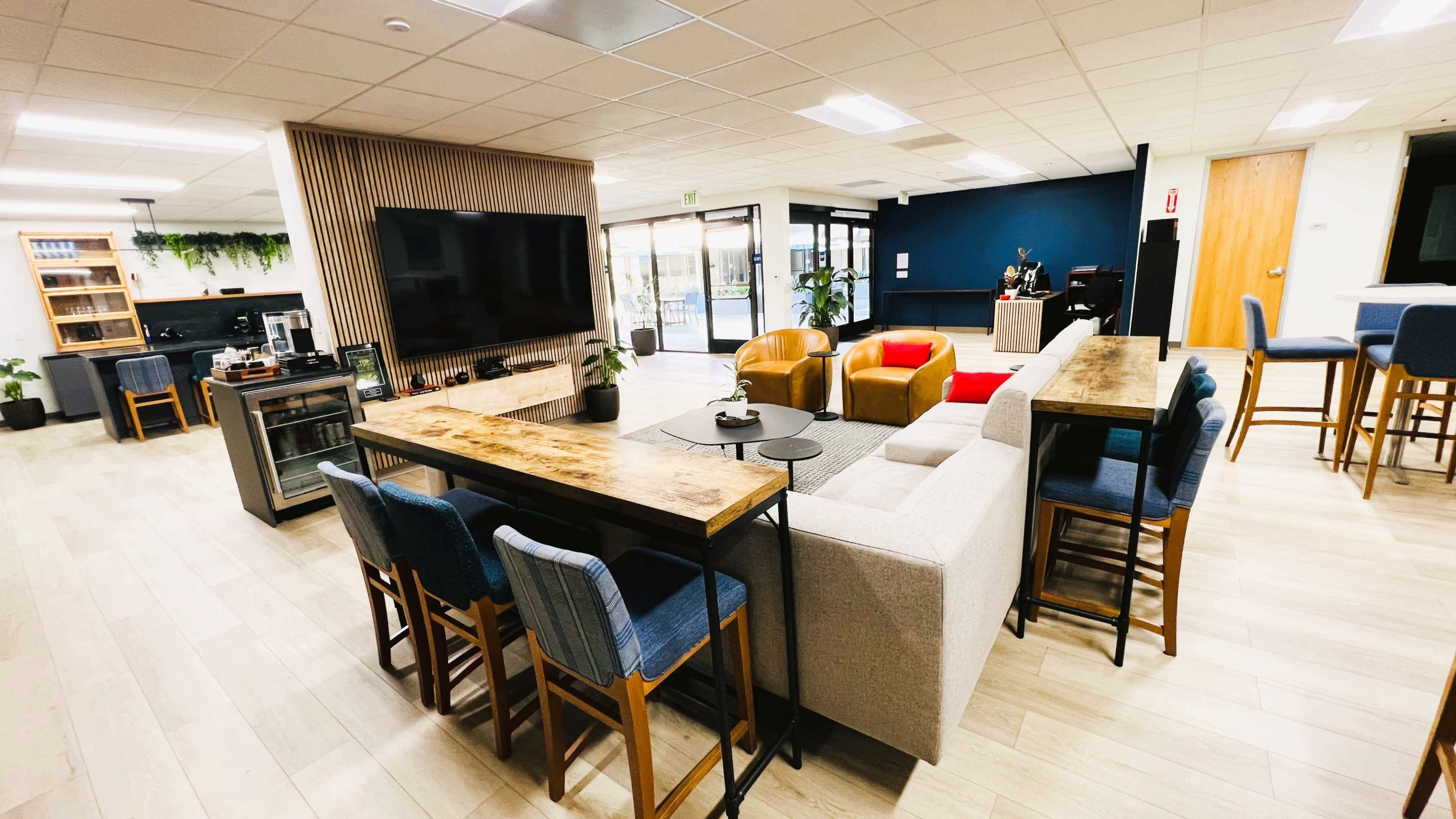 The image shows a modern communal lounge area with a mix of seating, including a long table with bar stools, a gray couch, and two armchairs, alongside a large TV and various plants.