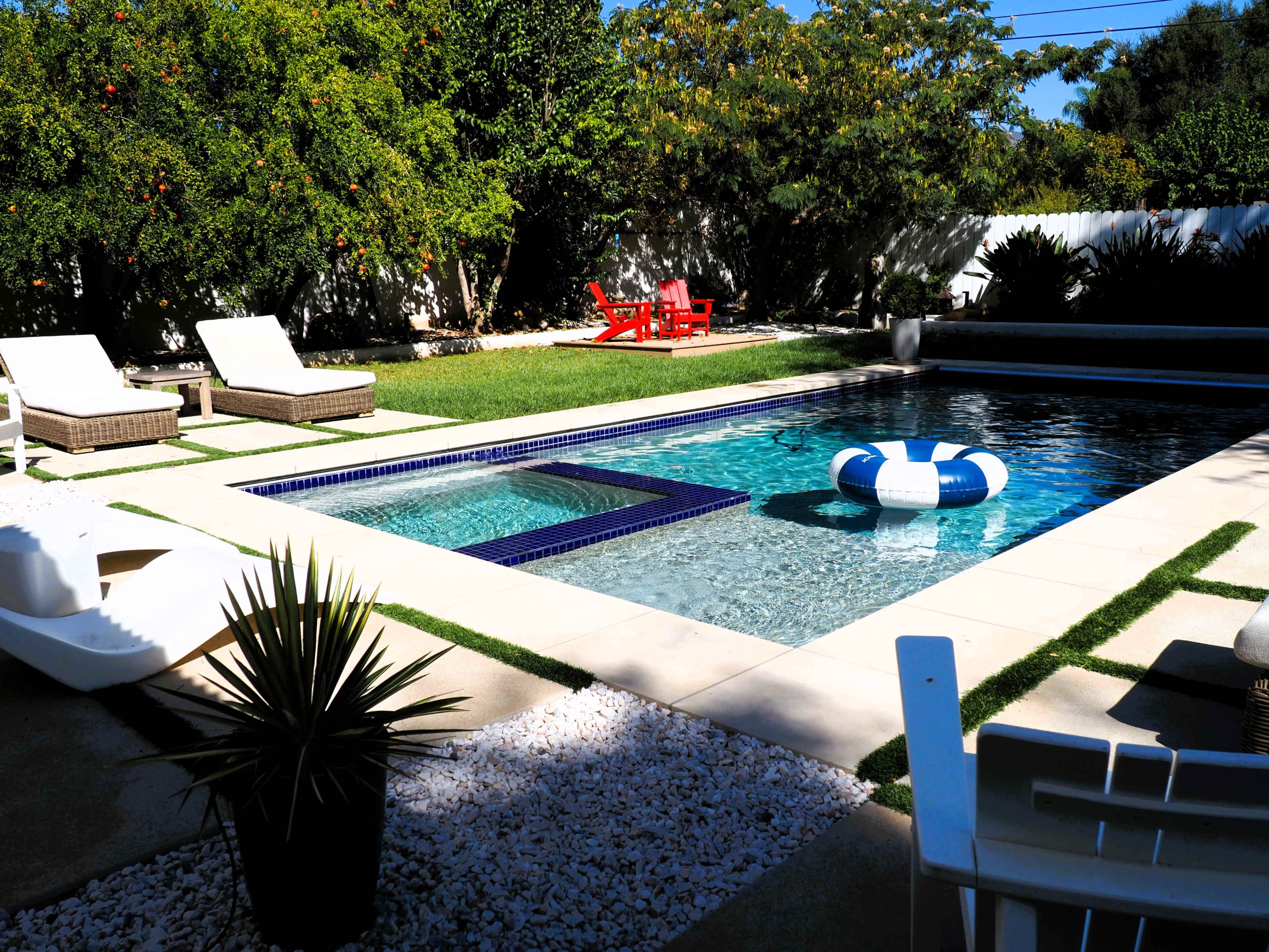 The image shows a landscaped backyard with a swimming pool, lounge chairs, and a red outdoor table and chairs under a blue sky.