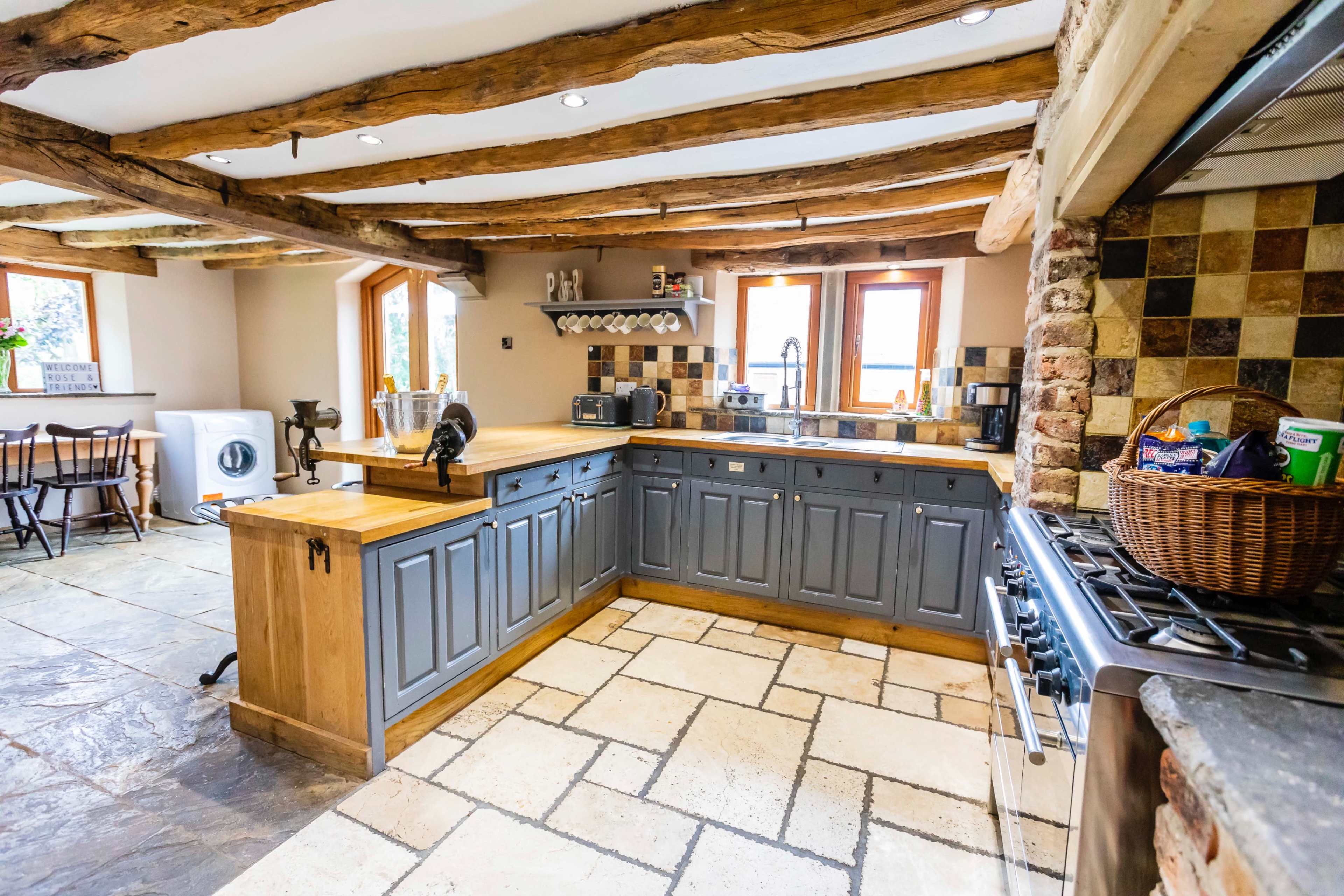 The kitchen features a wooden beam ceiling, gray cabinetry, a large wooden countertop, and a stone tile floor.