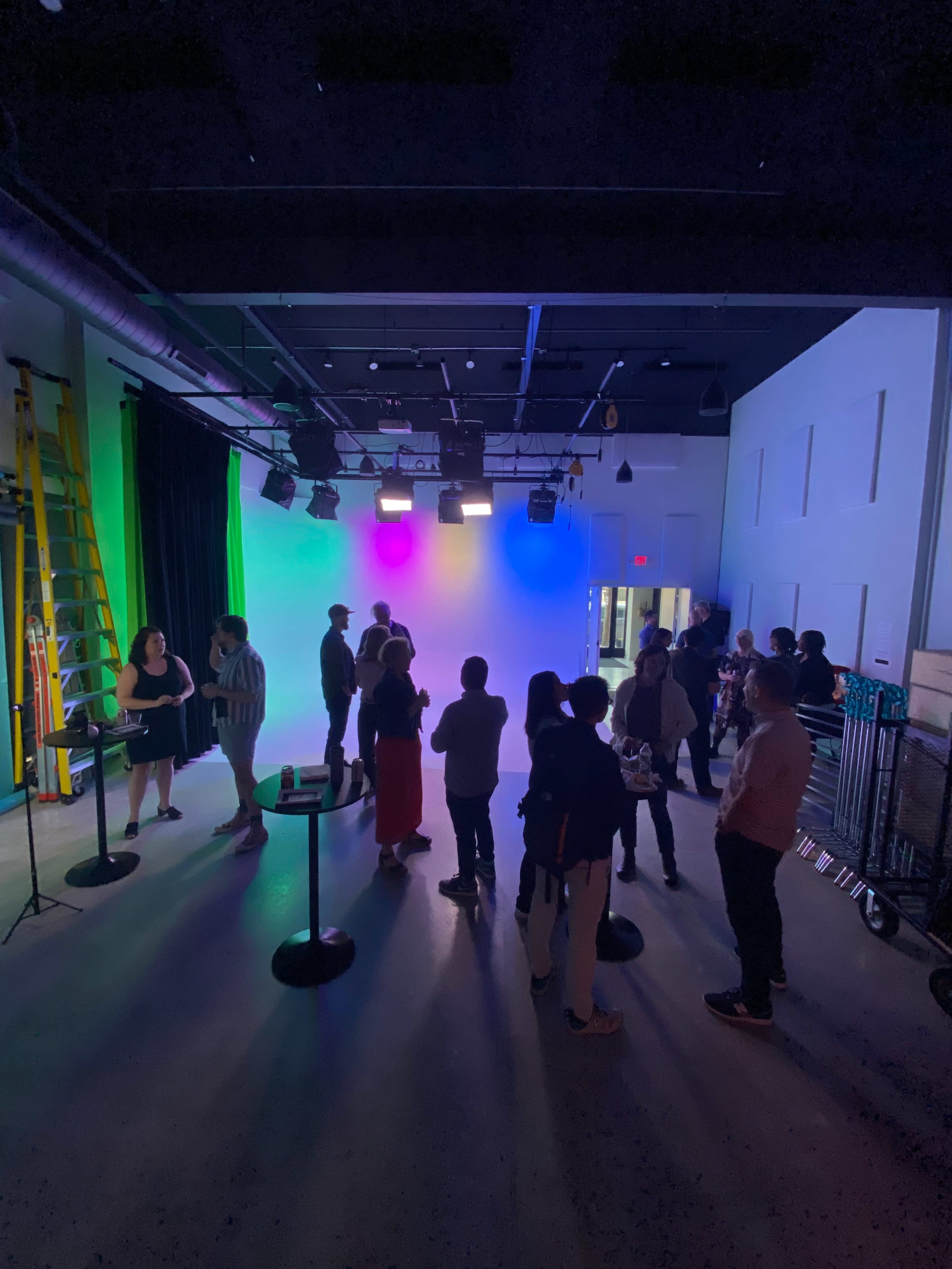 A group of people gathers in a brightly lit indoor space with colored backdrop lighting and various decorations.