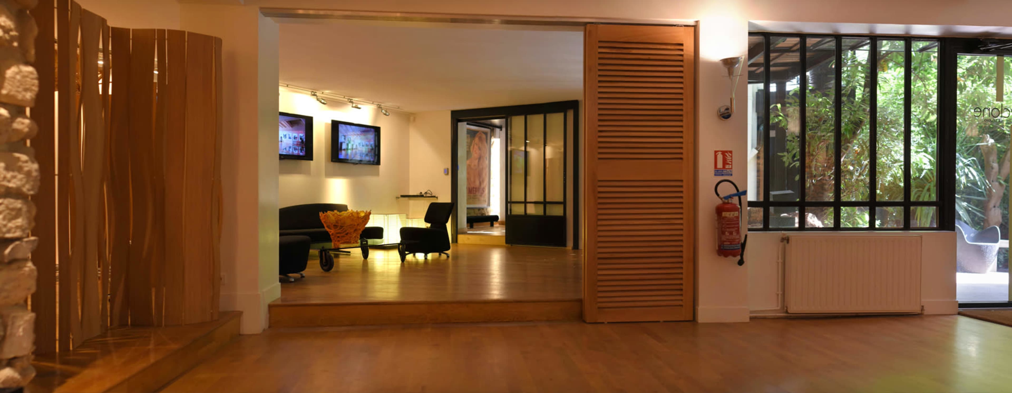 A spacious interior with wooden flooring, a hallway leading to a room with two screens, and large windows allowing natural light and a view of greenery.