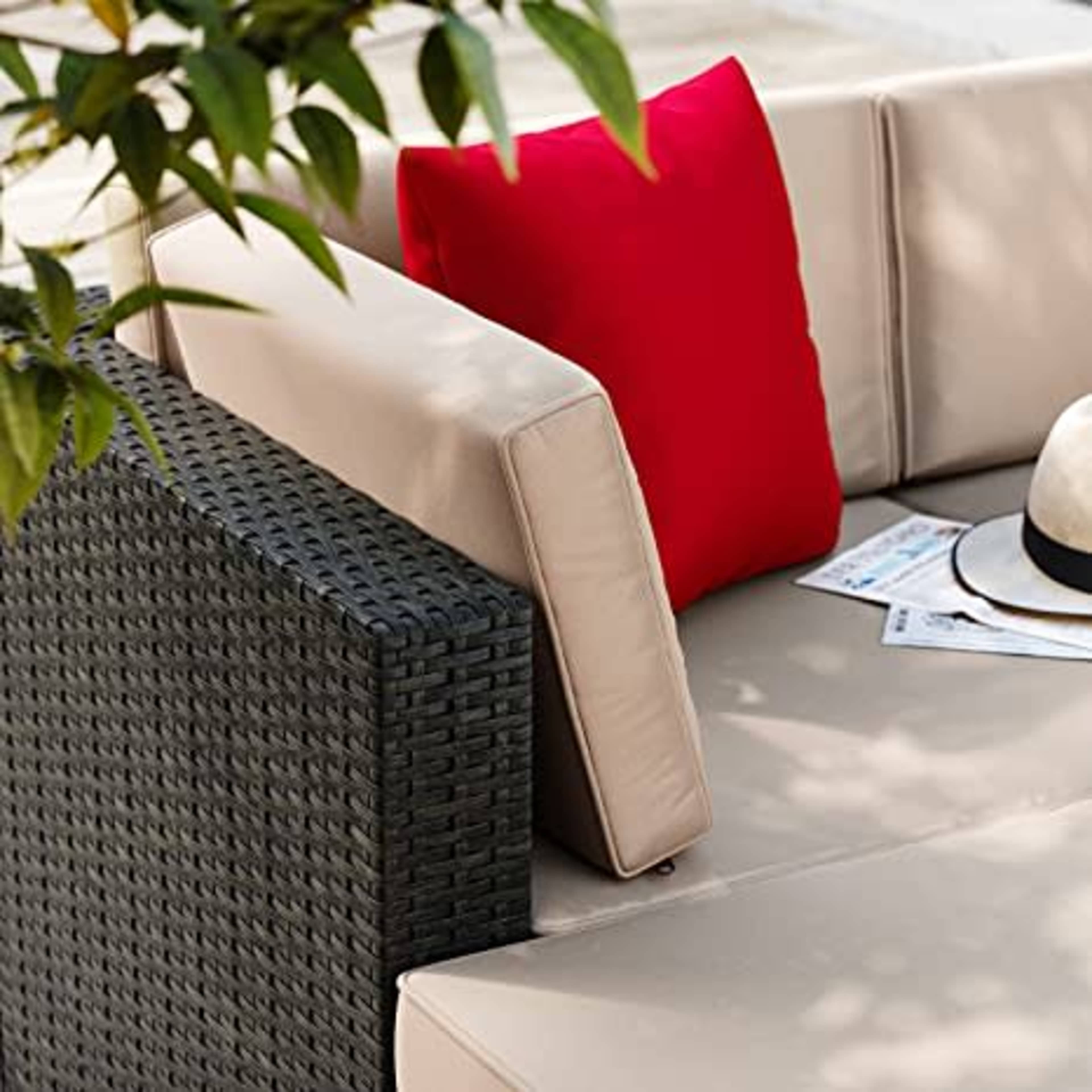 The image shows a corner of an outdoor sofa featuring beige cushions, a red decorative pillow, and a textured black armrest, with a straw hat and some newspapers nearby.