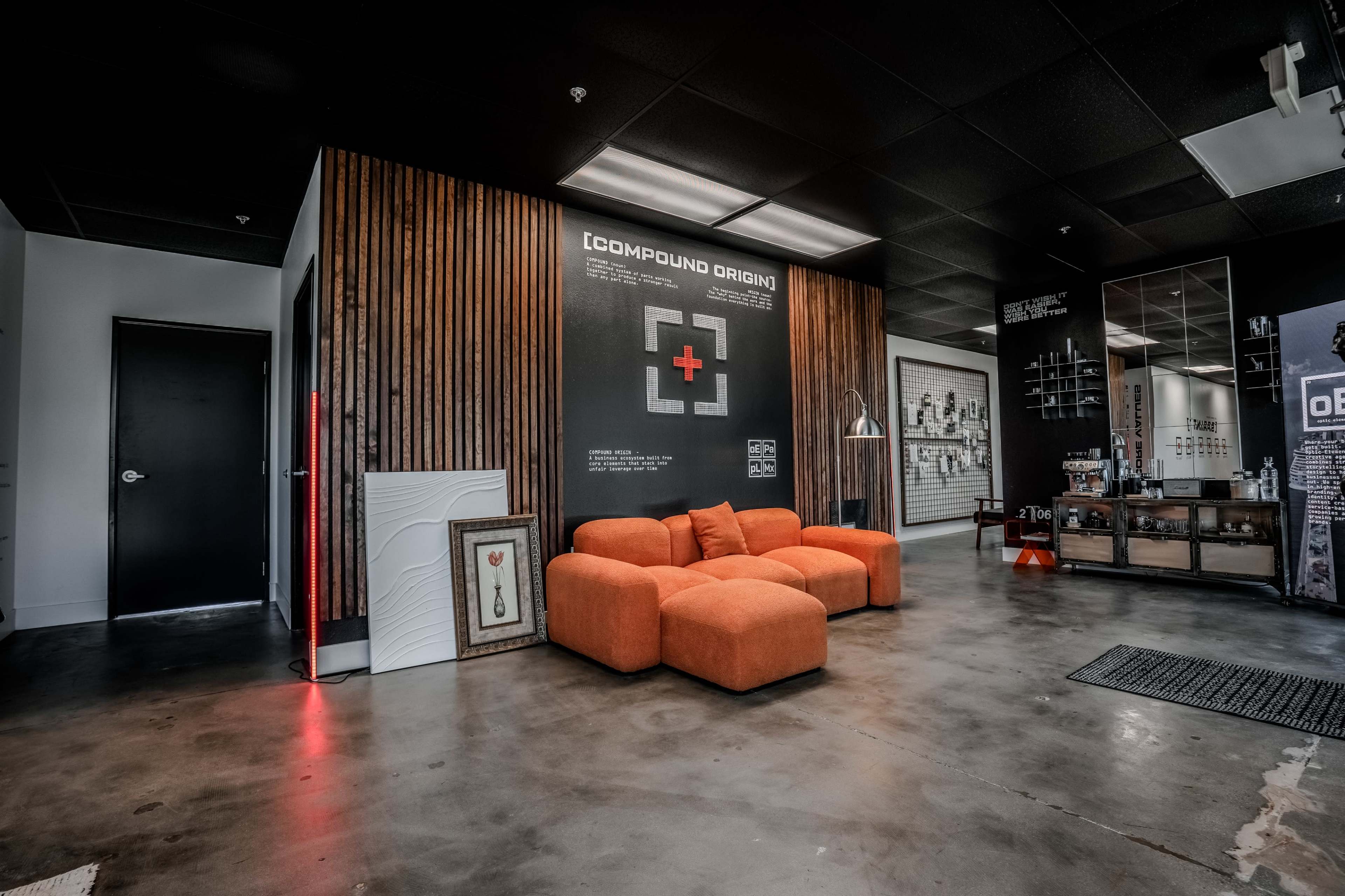 The image shows a modern office interior featuring an orange sectional sofa, a wooden accent wall, and various artworks.