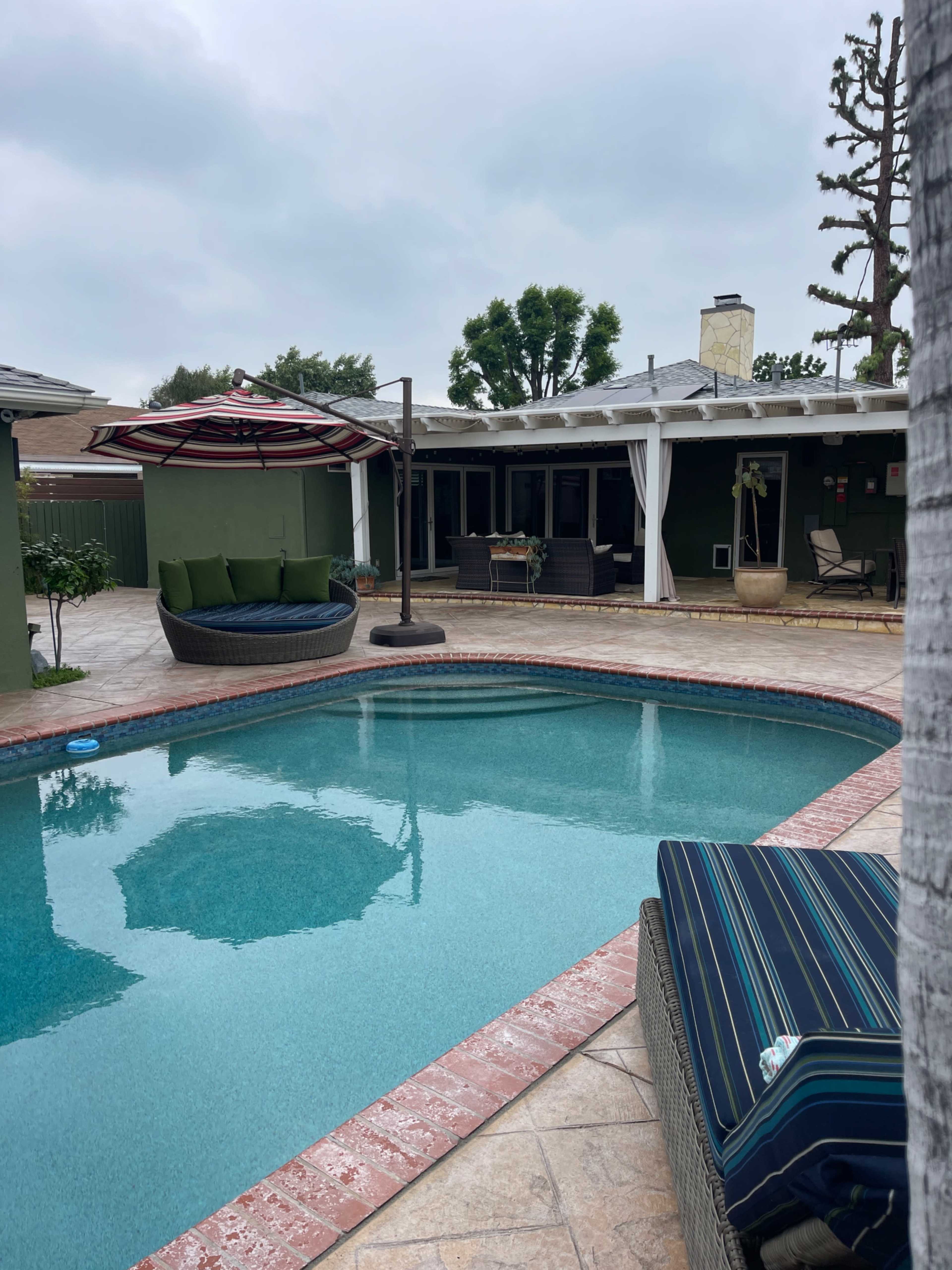 A backyard pool area with a round umbrella, lounge chairs, and a shaded seating area next to a house.