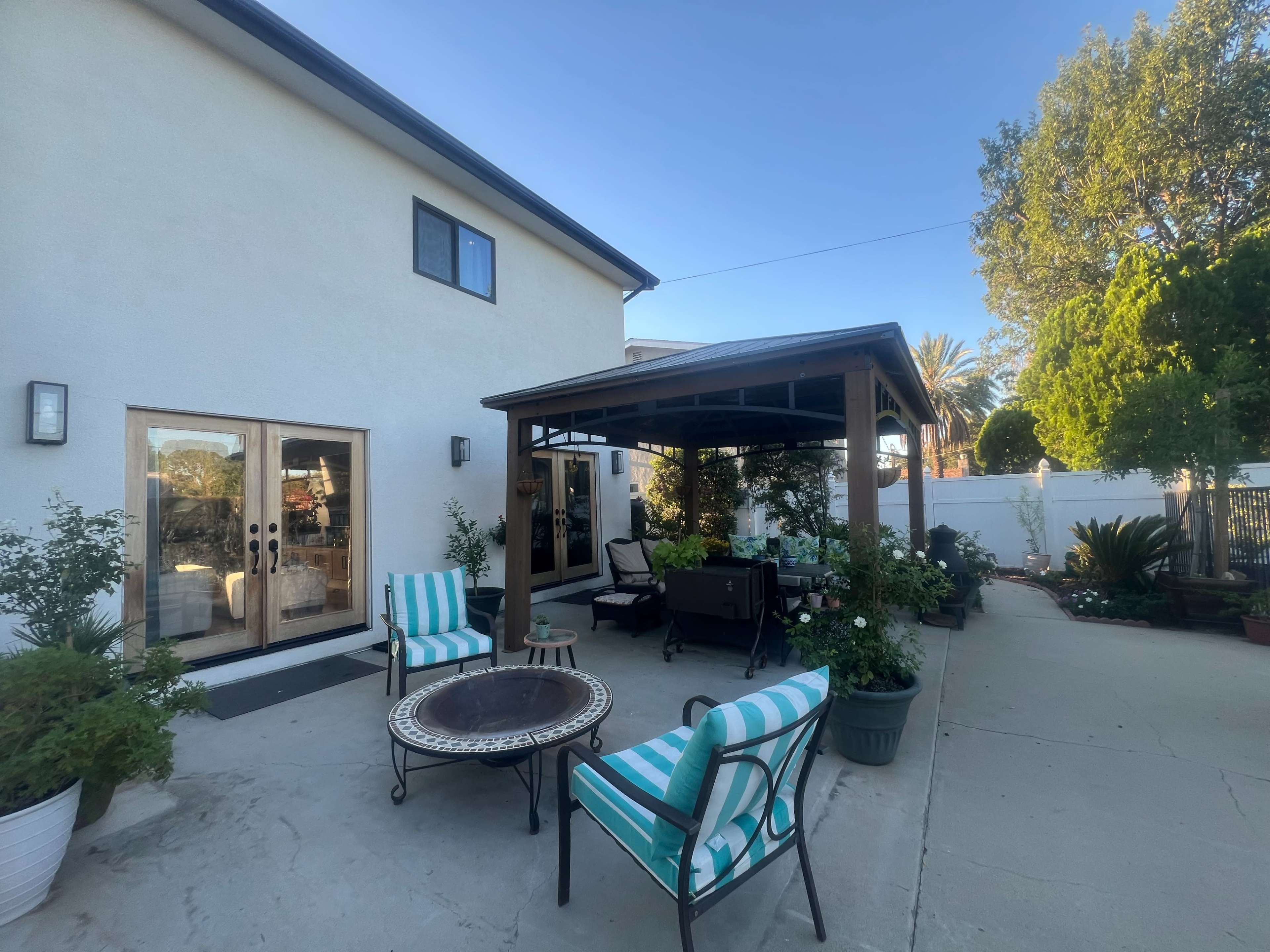 A backyard area features a covered patio with seating, a fire pit, and surrounding plants and trees.