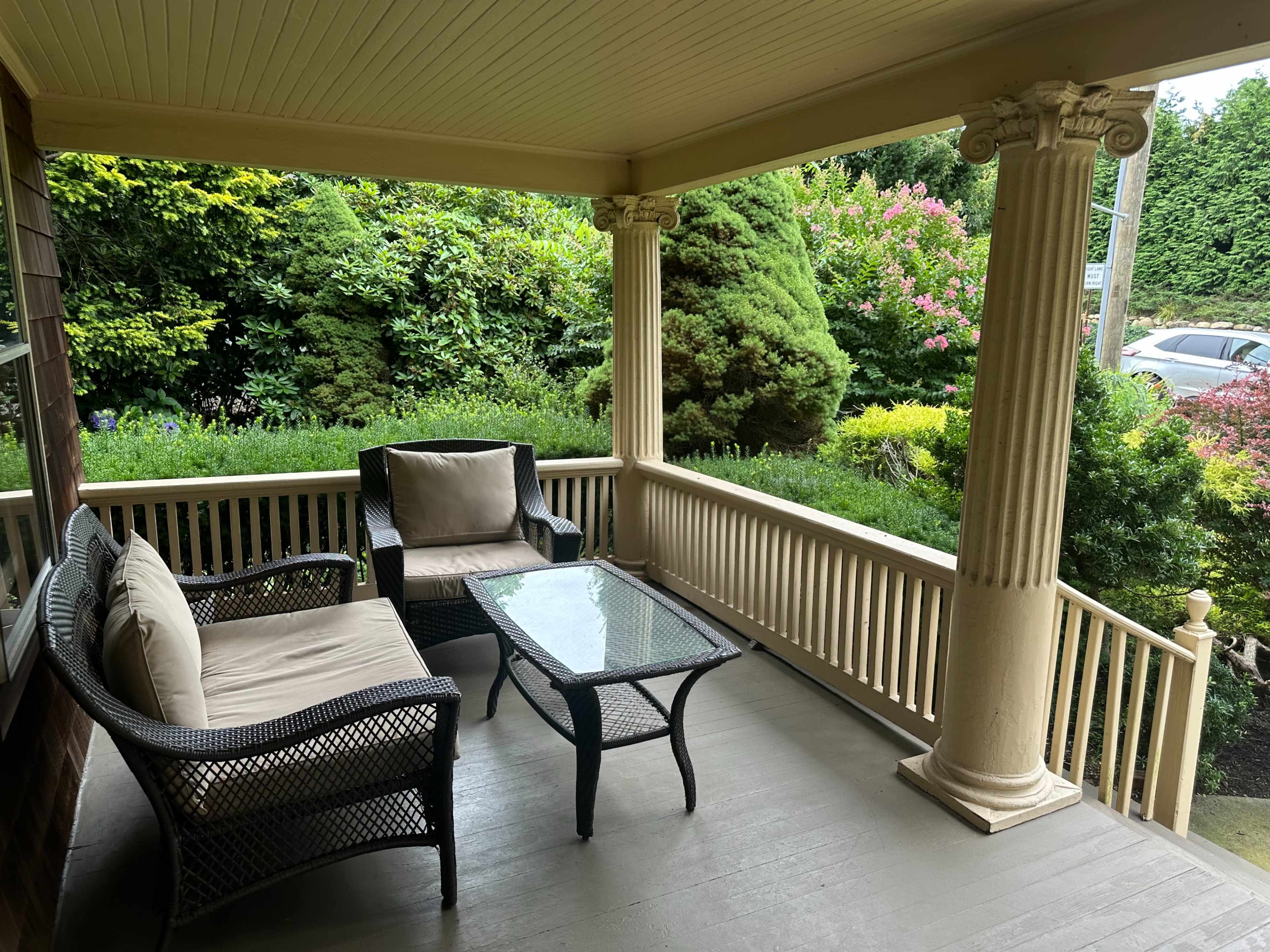 A porch includes a glass coffee table and two wicker chairs with cushions, surrounded by lush greenery and colorful plants.