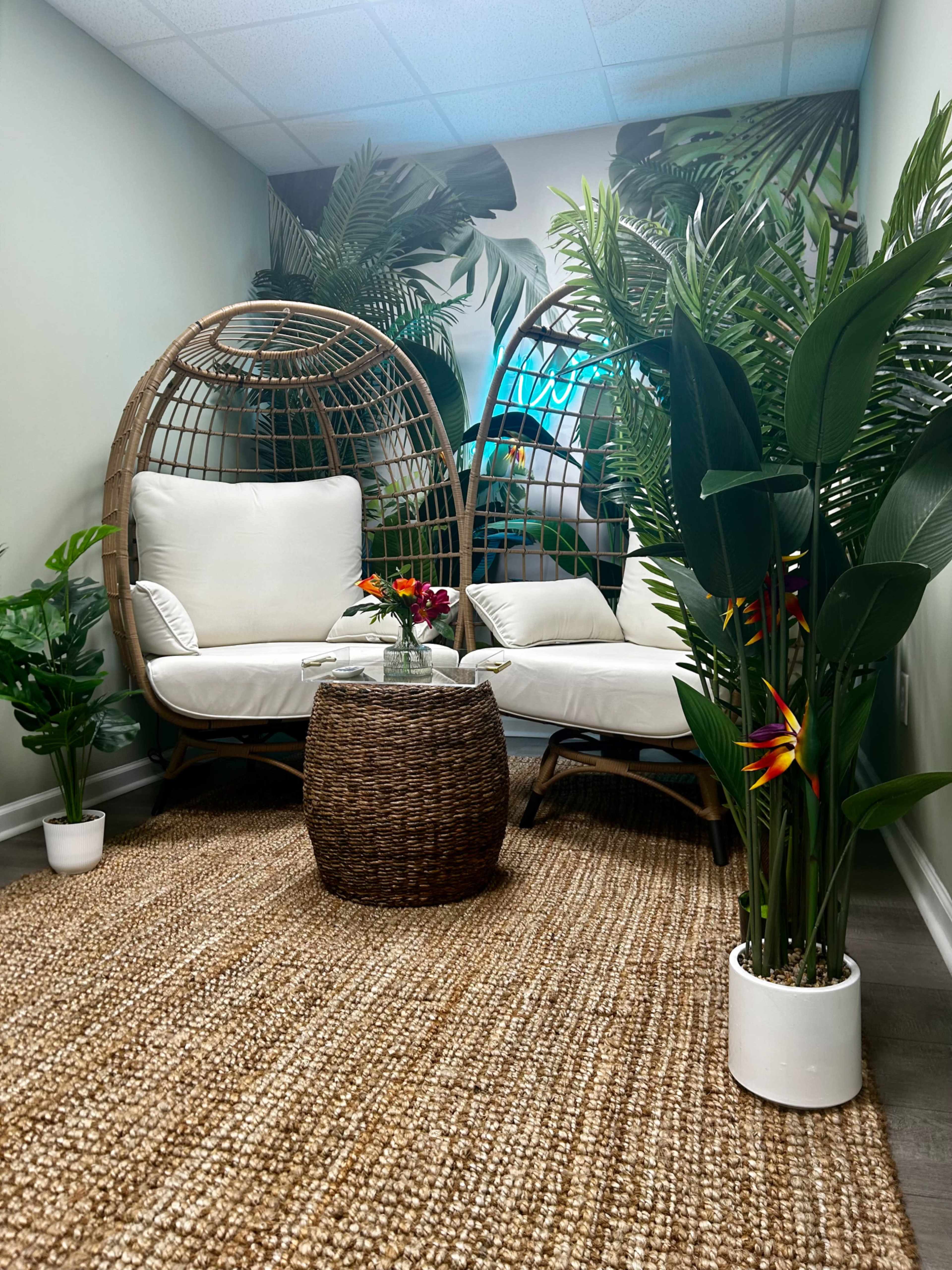 The image shows a cozy seating area featuring two wicker egg chairs, surrounded by lush green plants and a decorative round table at the center, all placed on a textured rug.