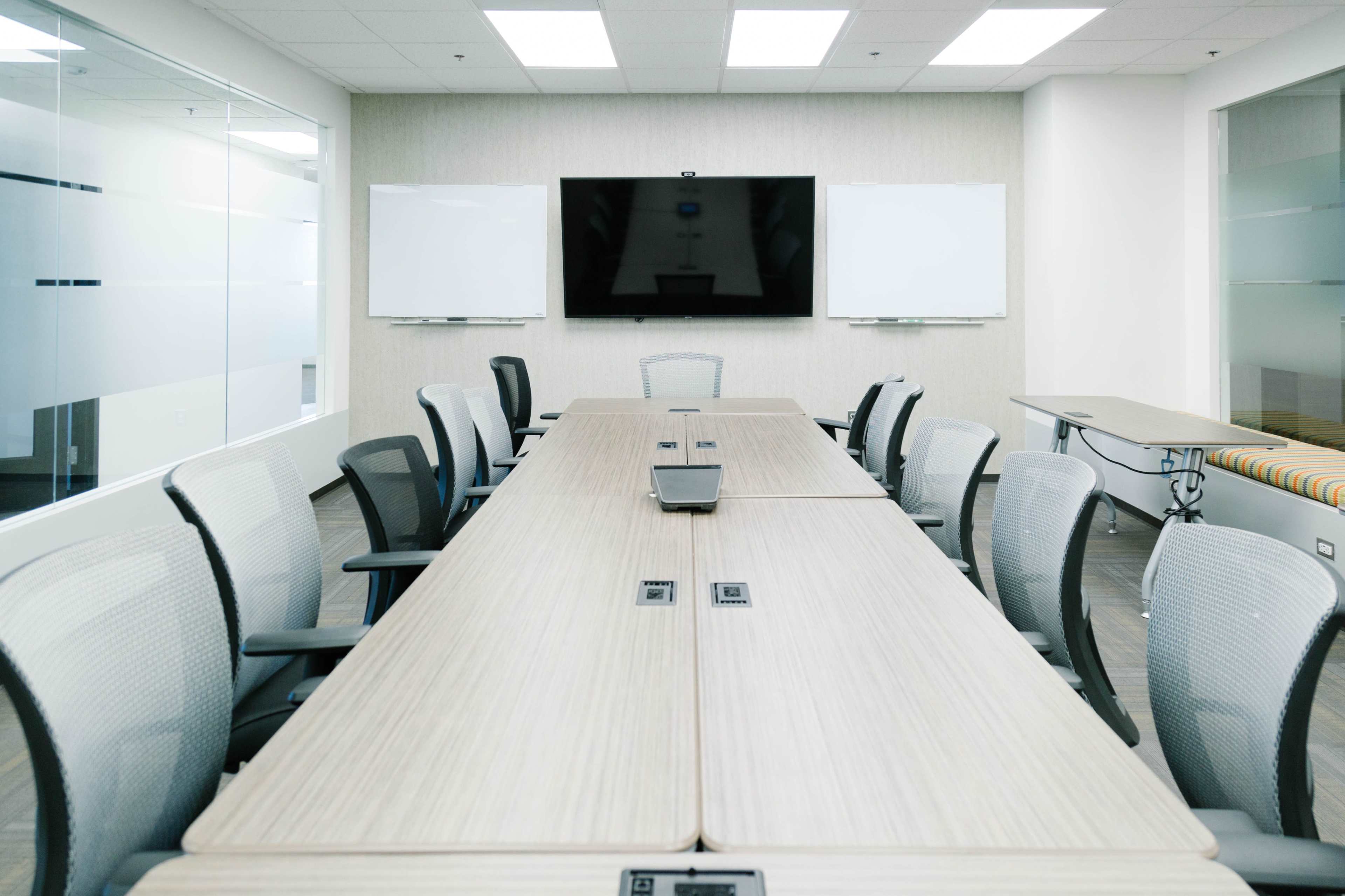 A modern conference room features a long wooden table, multiple office chairs, and a large wall-mounted screen with whiteboards on either side.