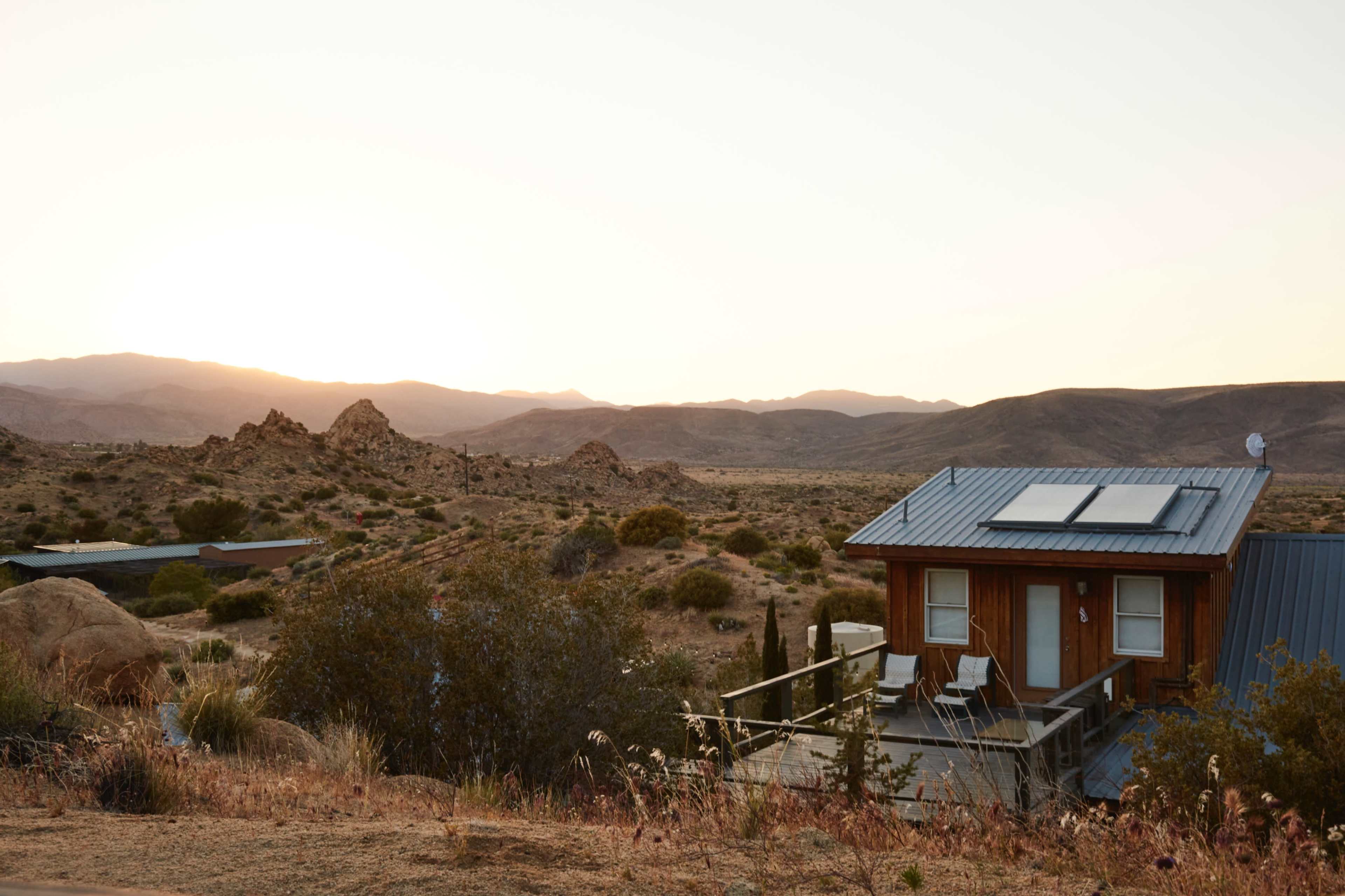 A small wooden house with a metal roof sits on a hillside overlooking a desert landscape at sunset.